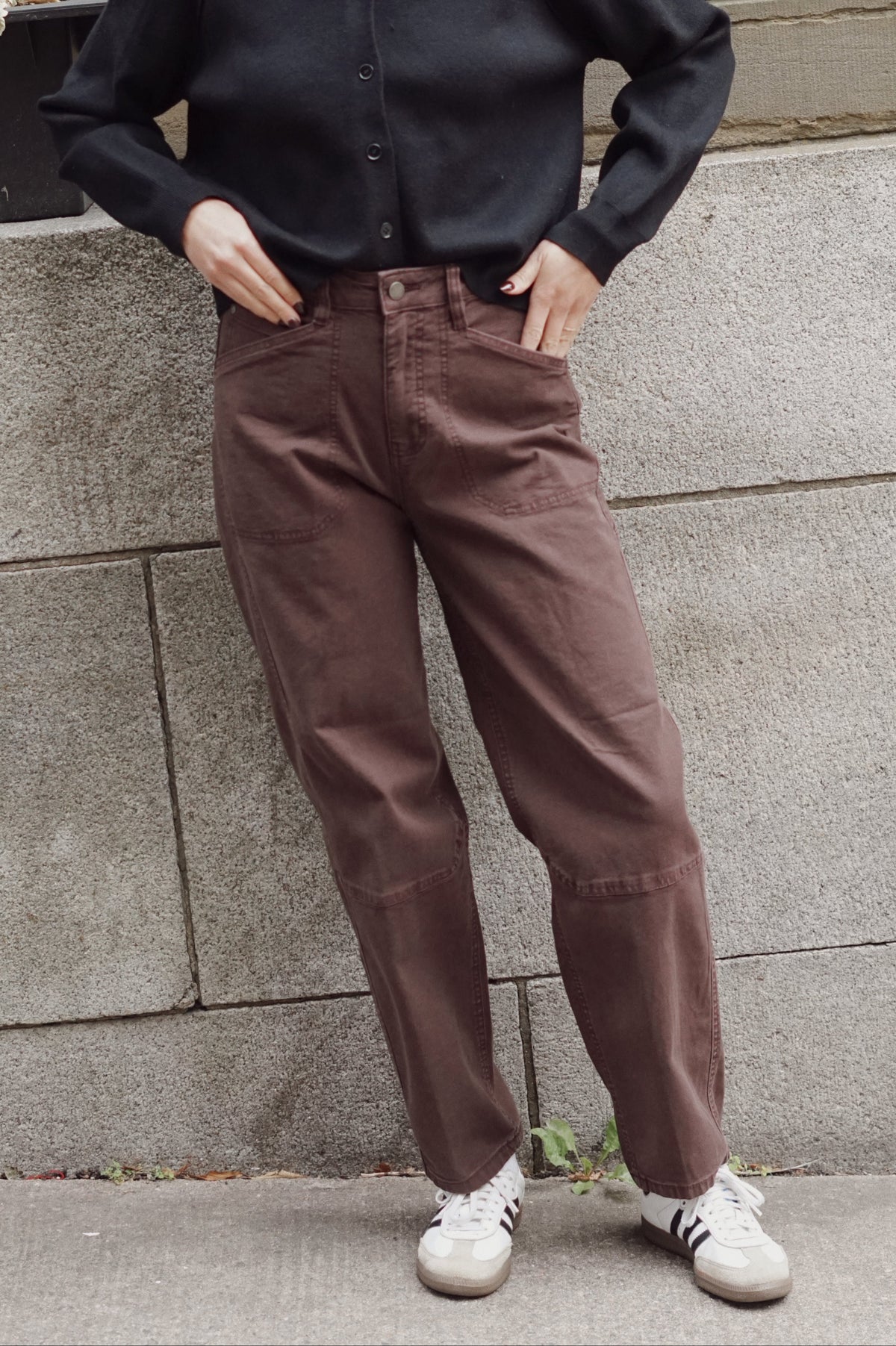 A person wearing brown wide-leg utility pants, a black long-sleeve button-up shirt, and white sneakers, standing against a gray stone wall.