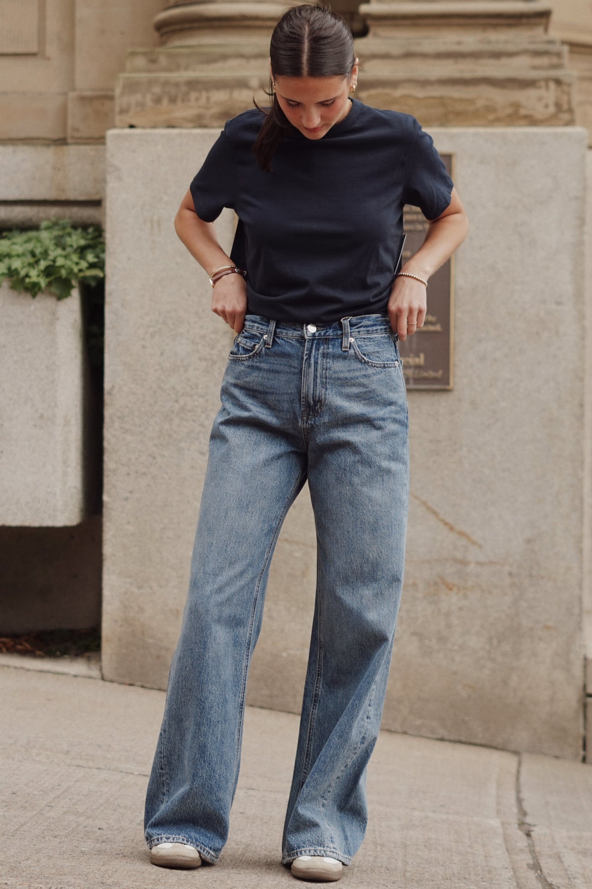 A woman wearing a dark navy t-shirt and light-wash blue wide-leg jeans, standing outdoors on a concrete sidewalk in front of a stone building.