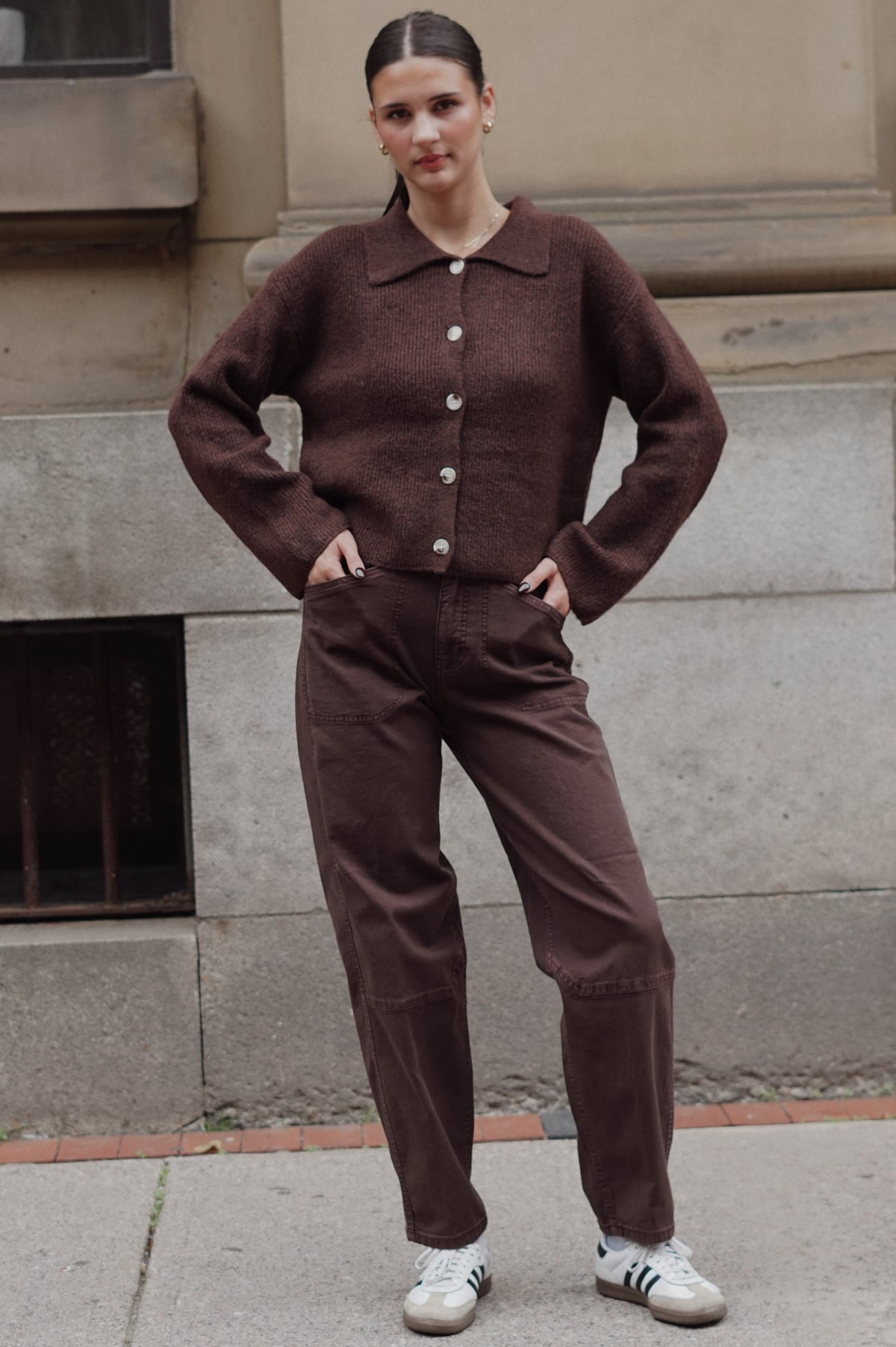 A woman stands outdoors against a stone building wall wearing a matching brown knit button-front cardigan and brown pants, with white sneakers.