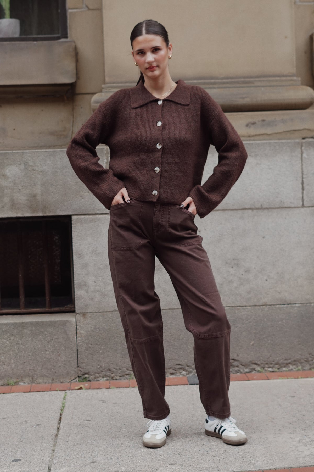 A woman stands outdoors against a stone building wall wearing a matching dark brown knit button-front cardigan and wide-leg brown pants, with white sneakers.