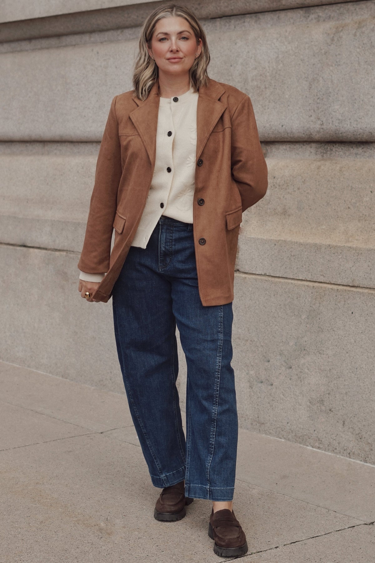 A woman wearing a brown suede-like blazer over a cream button-front top, blue jeans, and brown shoes.