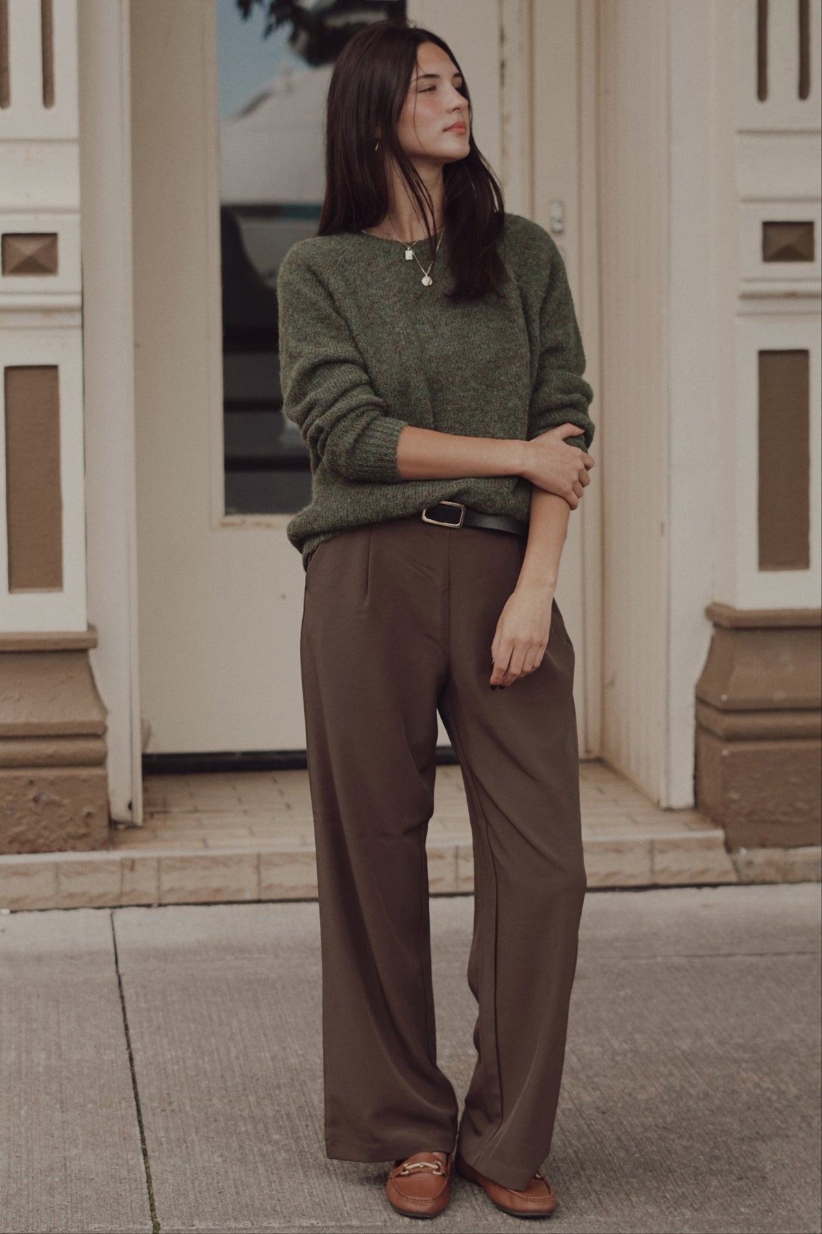 A woman wearing a textured olive green long-sleeve sweater, brown wide-leg trousers, and brown leather loafers.