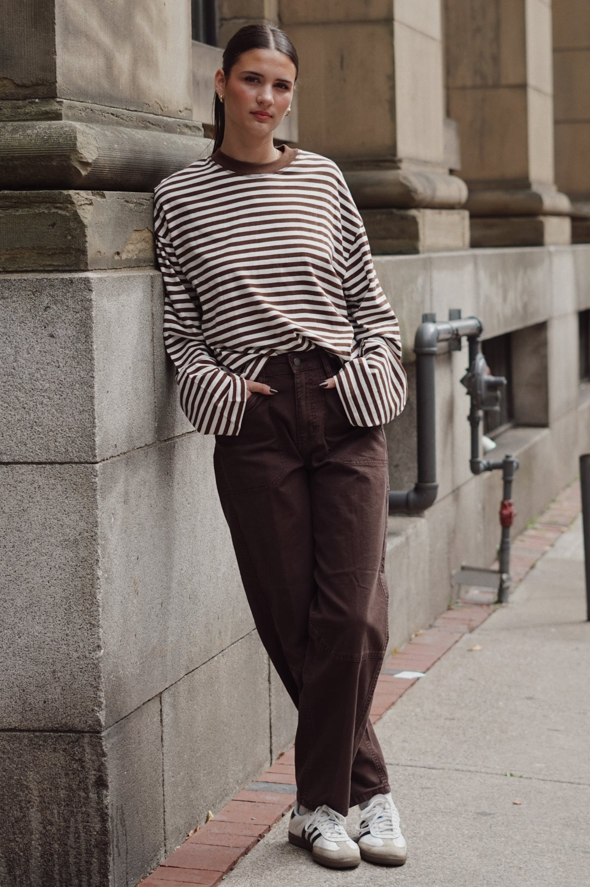 A woman stands on a city sidewalk leaning against a stone building, wearing a brown and cream horizontally striped long-sleeve top, dark brown pants, and white sneakers.