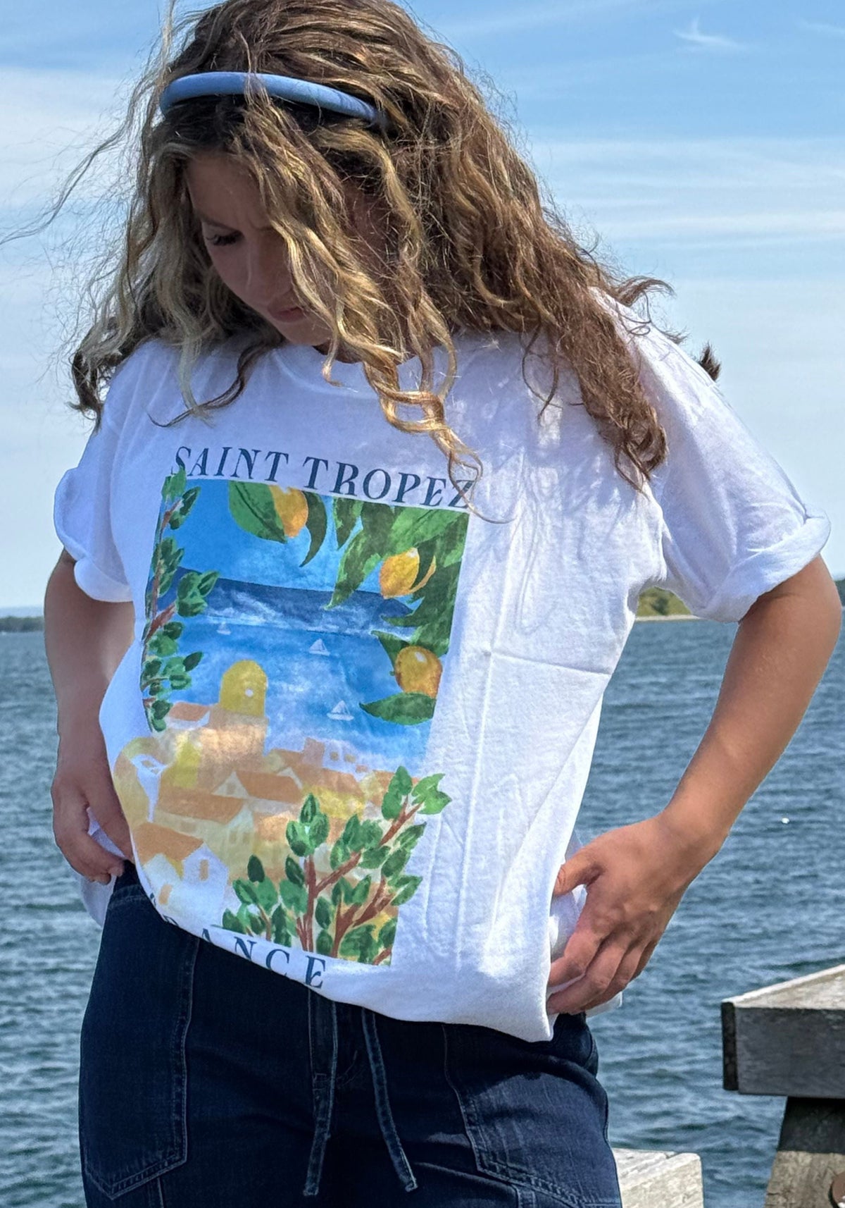 A person outdoors by the water wearing a white graphic T-shirt with 'SAINT TROPEZ' text and a colorful coastal scene print, paired with dark jeans.