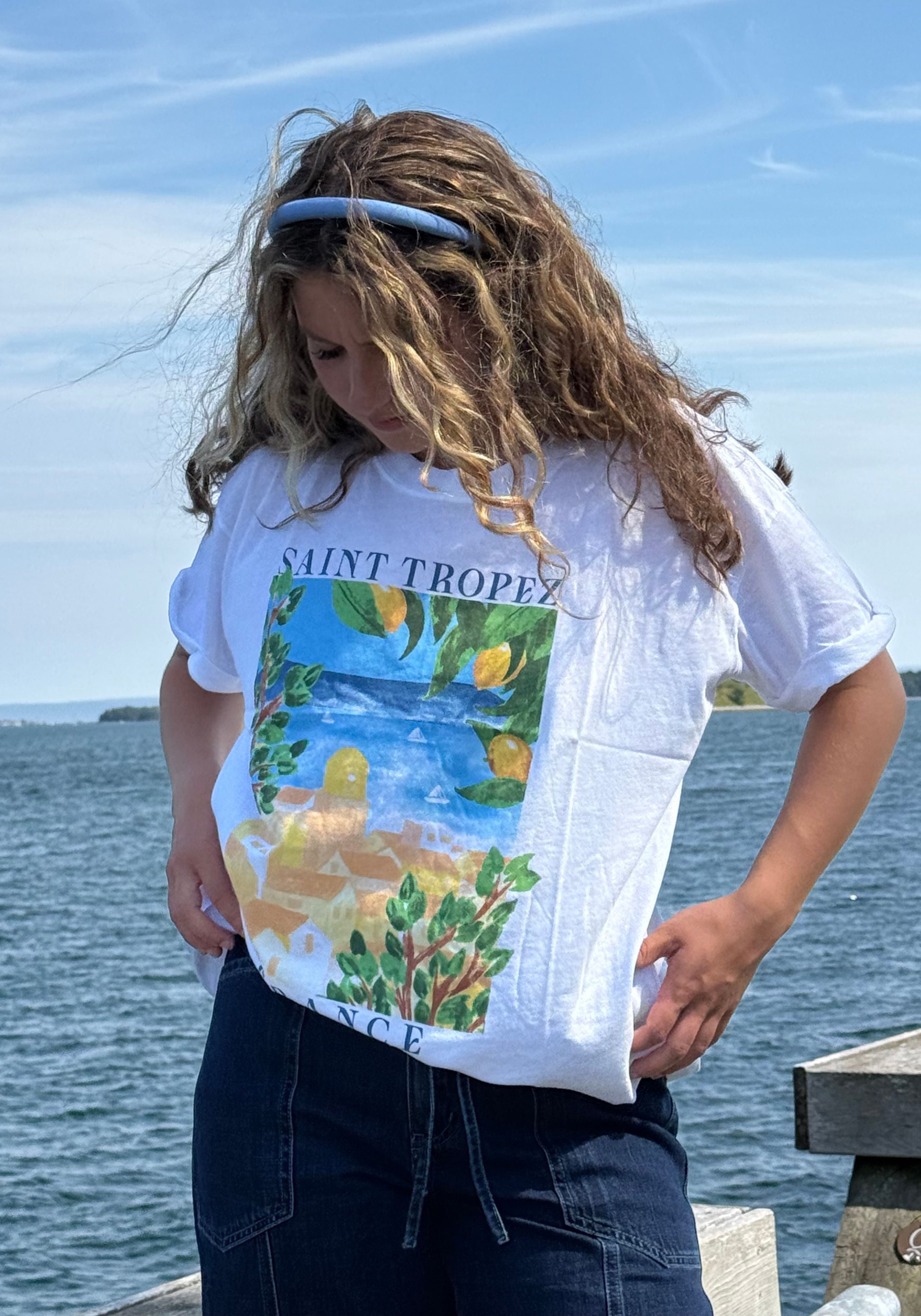 A person outdoors by the water wearing a white graphic T-shirt with 'SAINT TROPEZ' text and a colorful coastal scene print, paired with dark jeans.