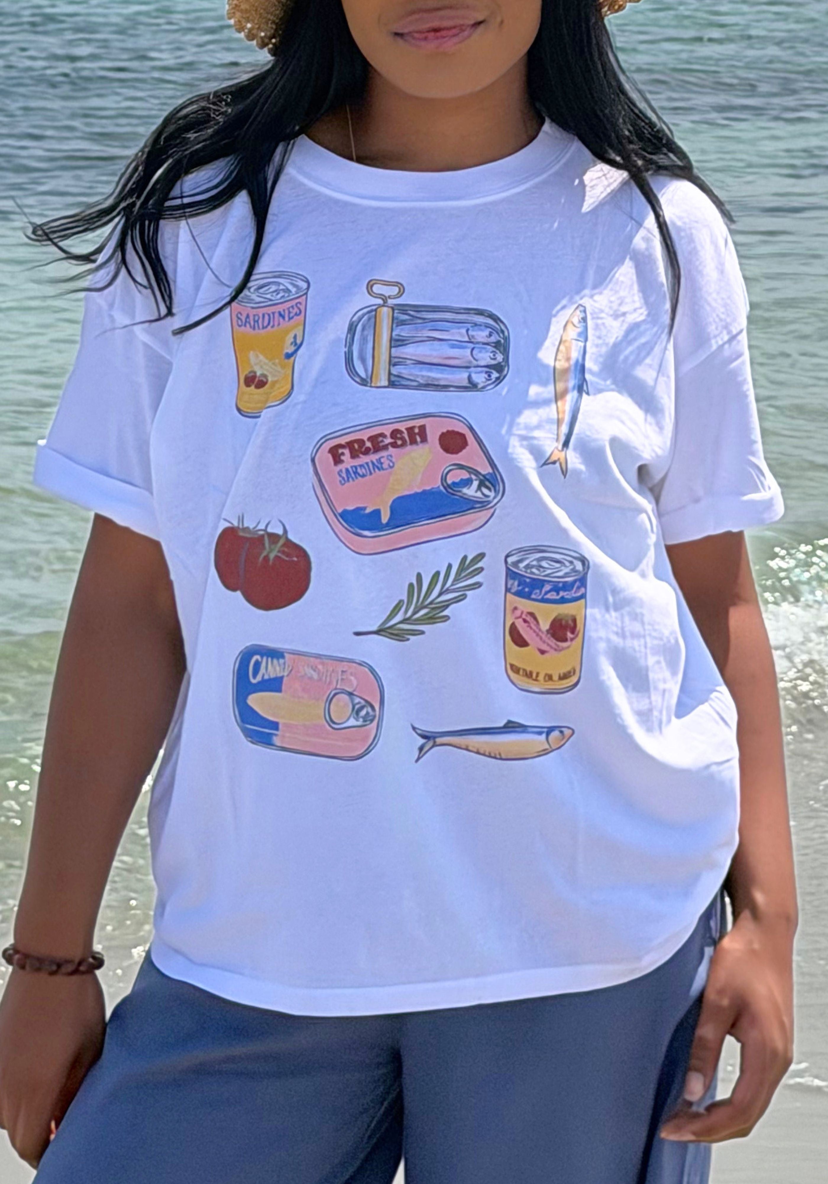 A woman wearing a white graphic t-shirt with colorful illustrations of canned fish, a tomato, and a green leaf, photographed outdoors near the ocean.