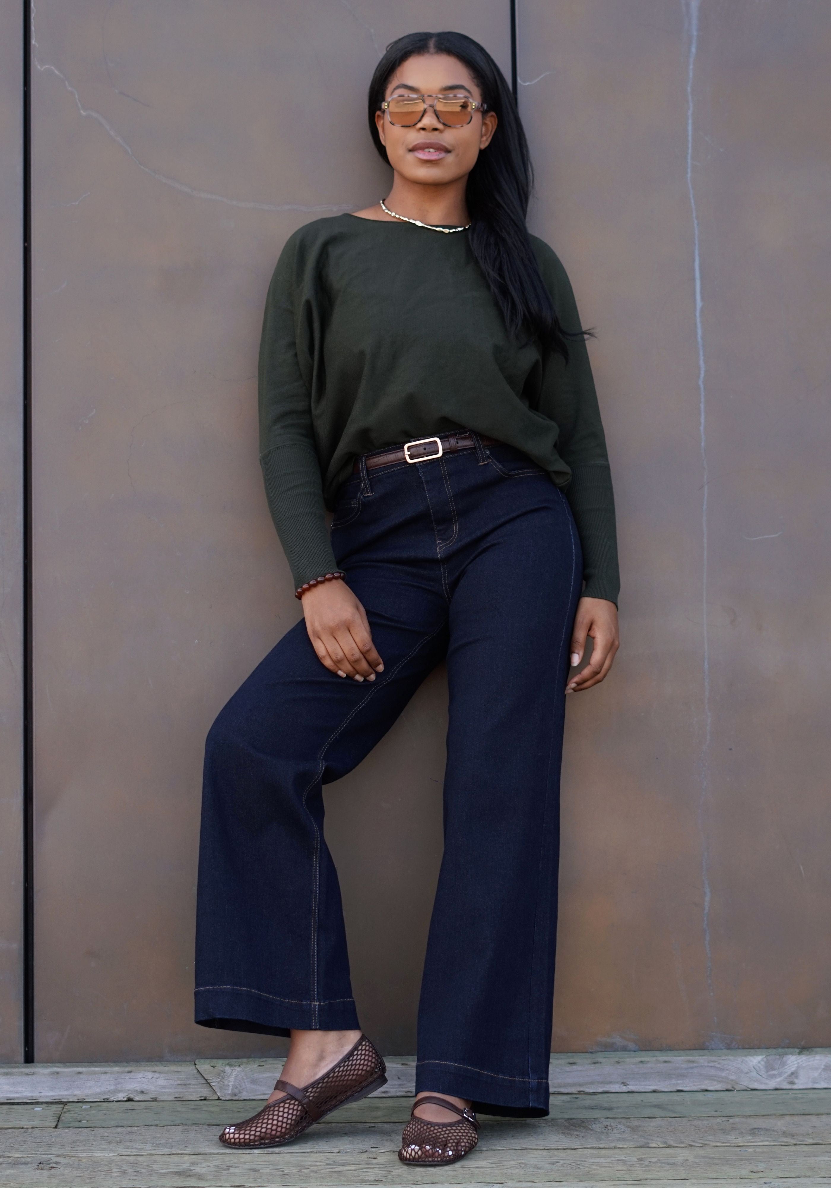 A woman wearing an olive green long-sleeve top, dark navy wide-leg jeans, brown flat shoes, and gold-framed sunglasses, leaning against a textured brown wall.