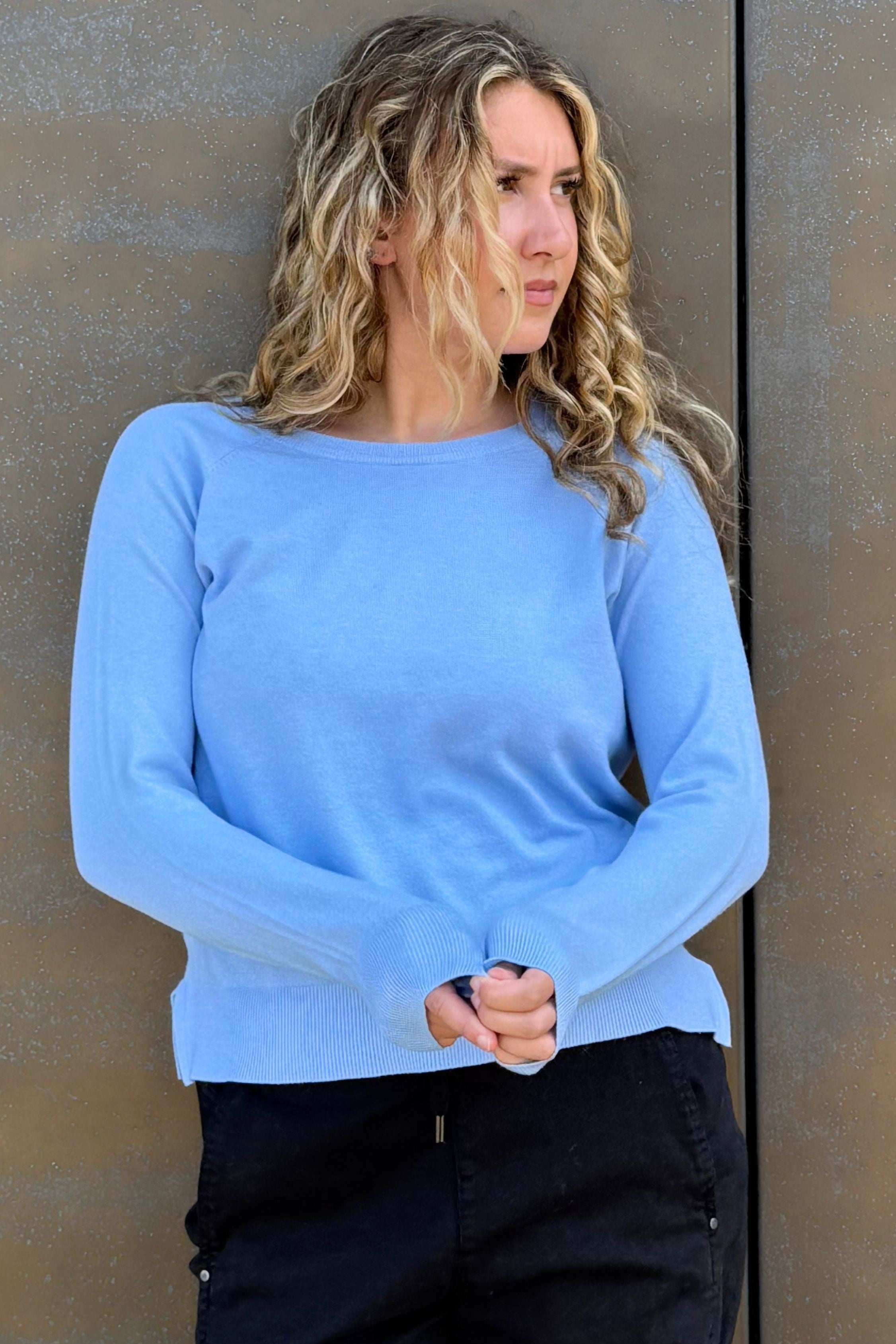 A woman wearing a light blue long-sleeve knit top and black pants, standing against a textured brown wall.