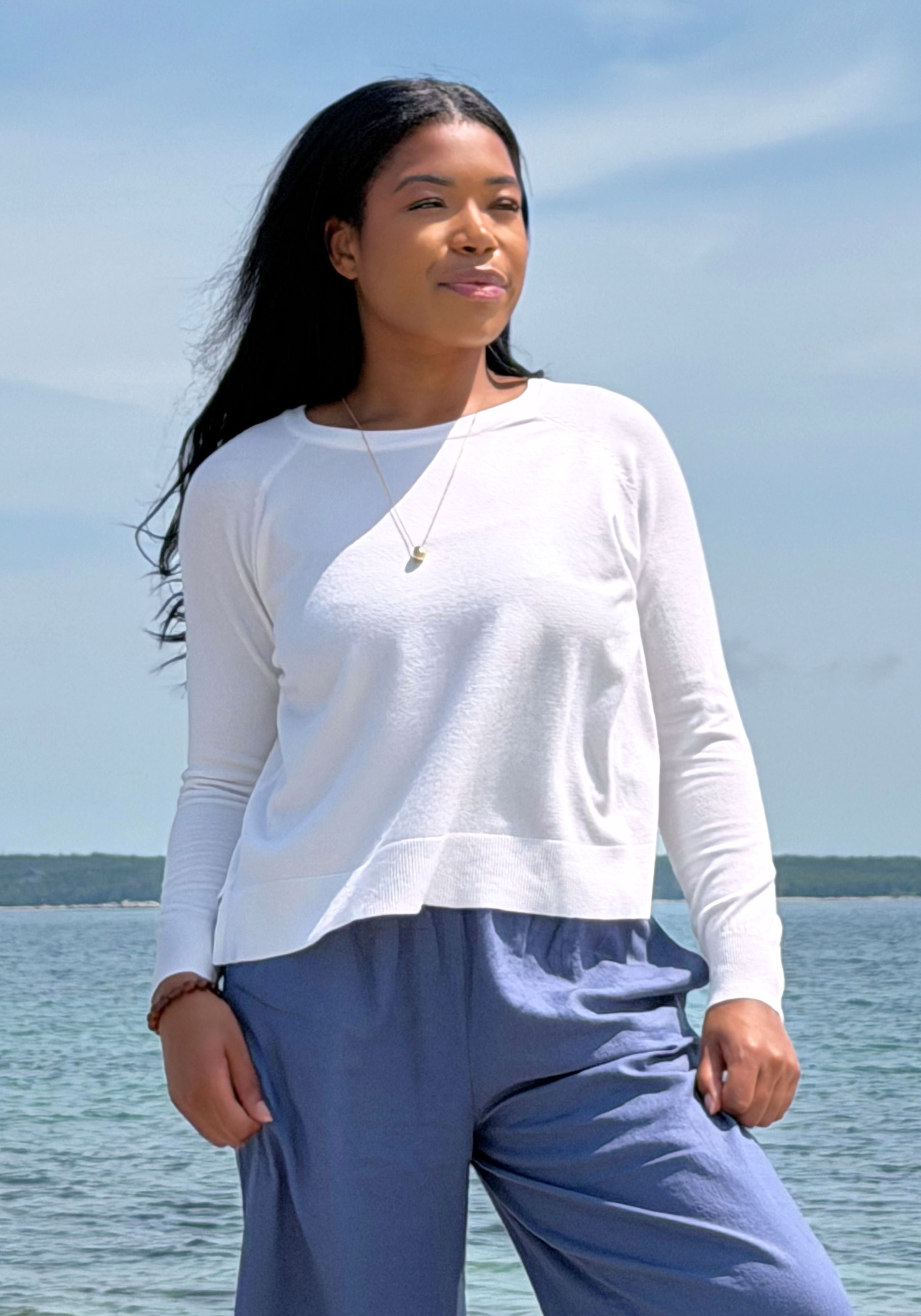 A woman wearing a white long-sleeve crewneck top and blue pants stands outdoors by the water under a blue sky.