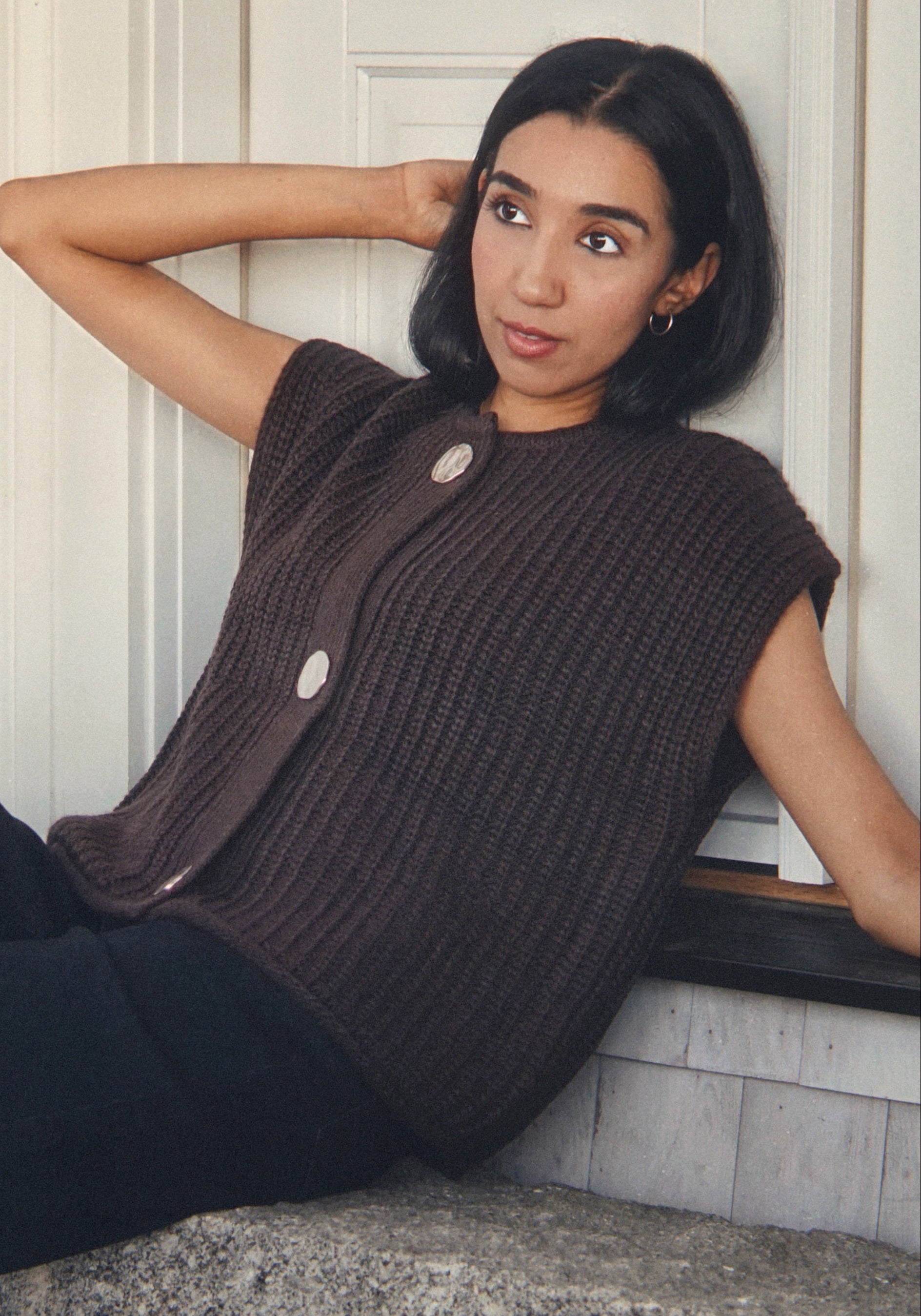 A woman sits on stone steps leaning against a white door, wearing a dark brown ribbed sleeveless cardigan with large buttons and black wide-leg pants.
