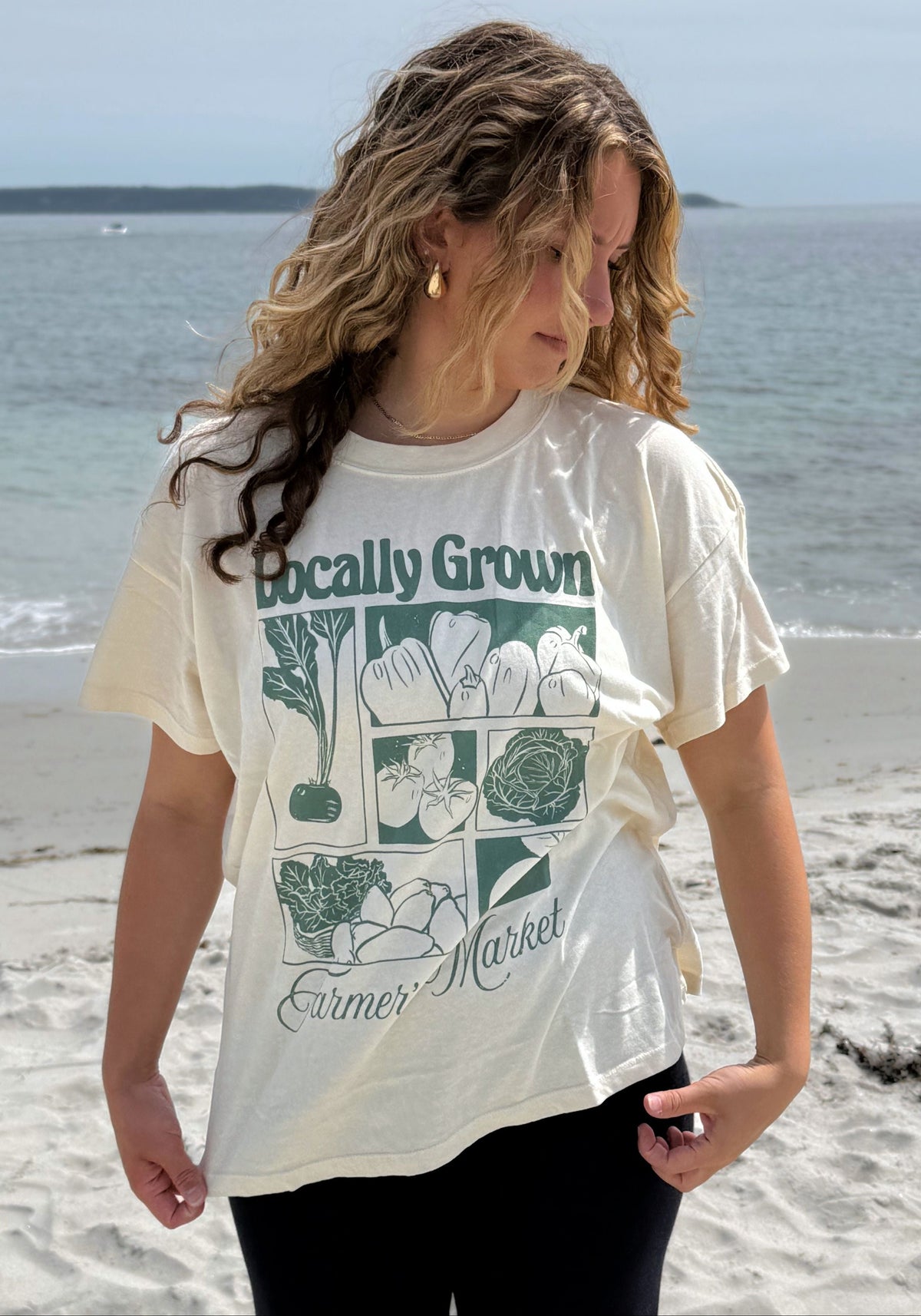 A woman standing on a sandy beach wearing a cream-colored graphic t-shirt with a green vegetable print and the text 'Locally Grown Farmer Market'.