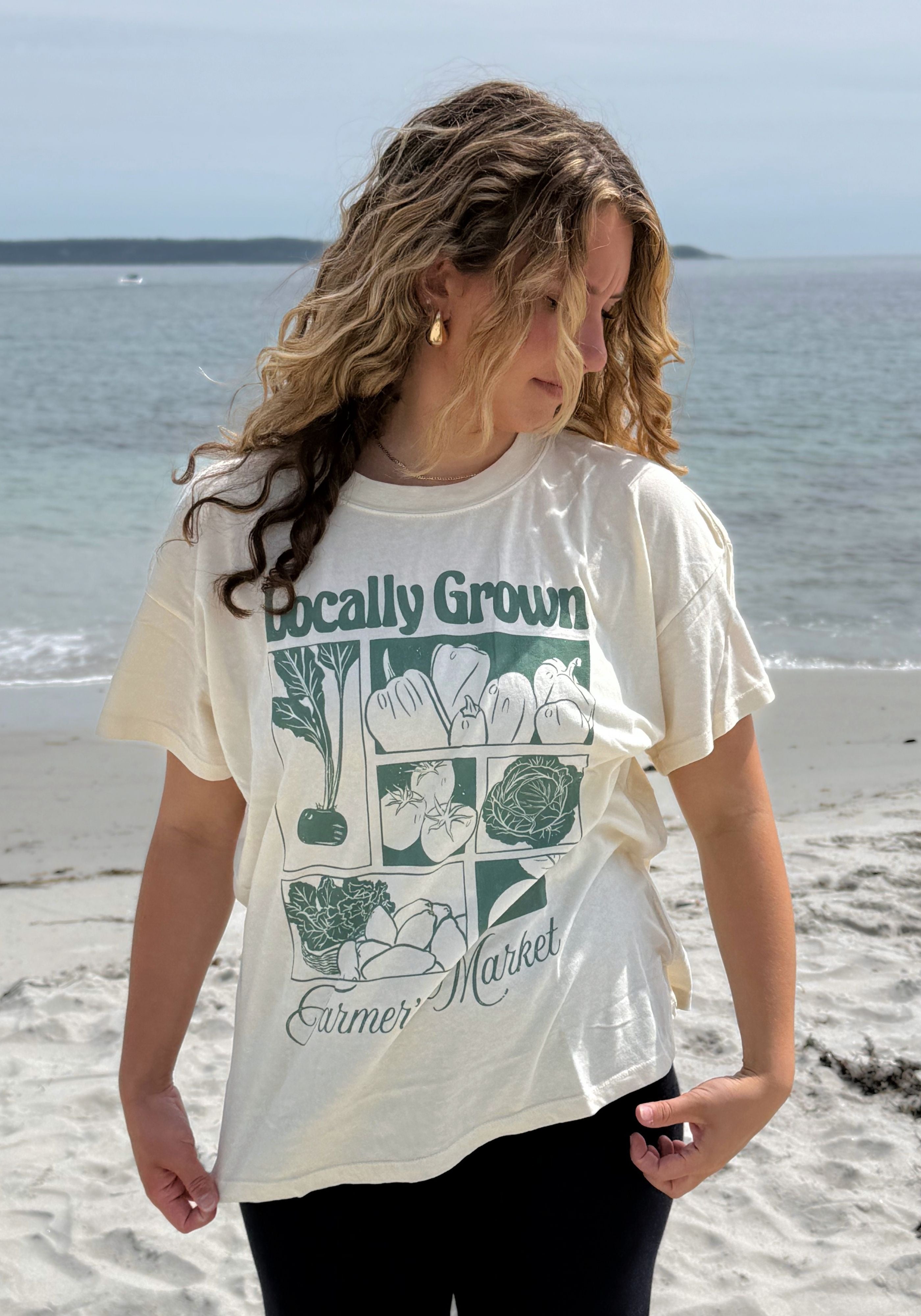 A woman standing on a sandy beach wearing a cream-colored graphic t-shirt with a green vegetable print and the text 'Locally Grown Farmer Market'.