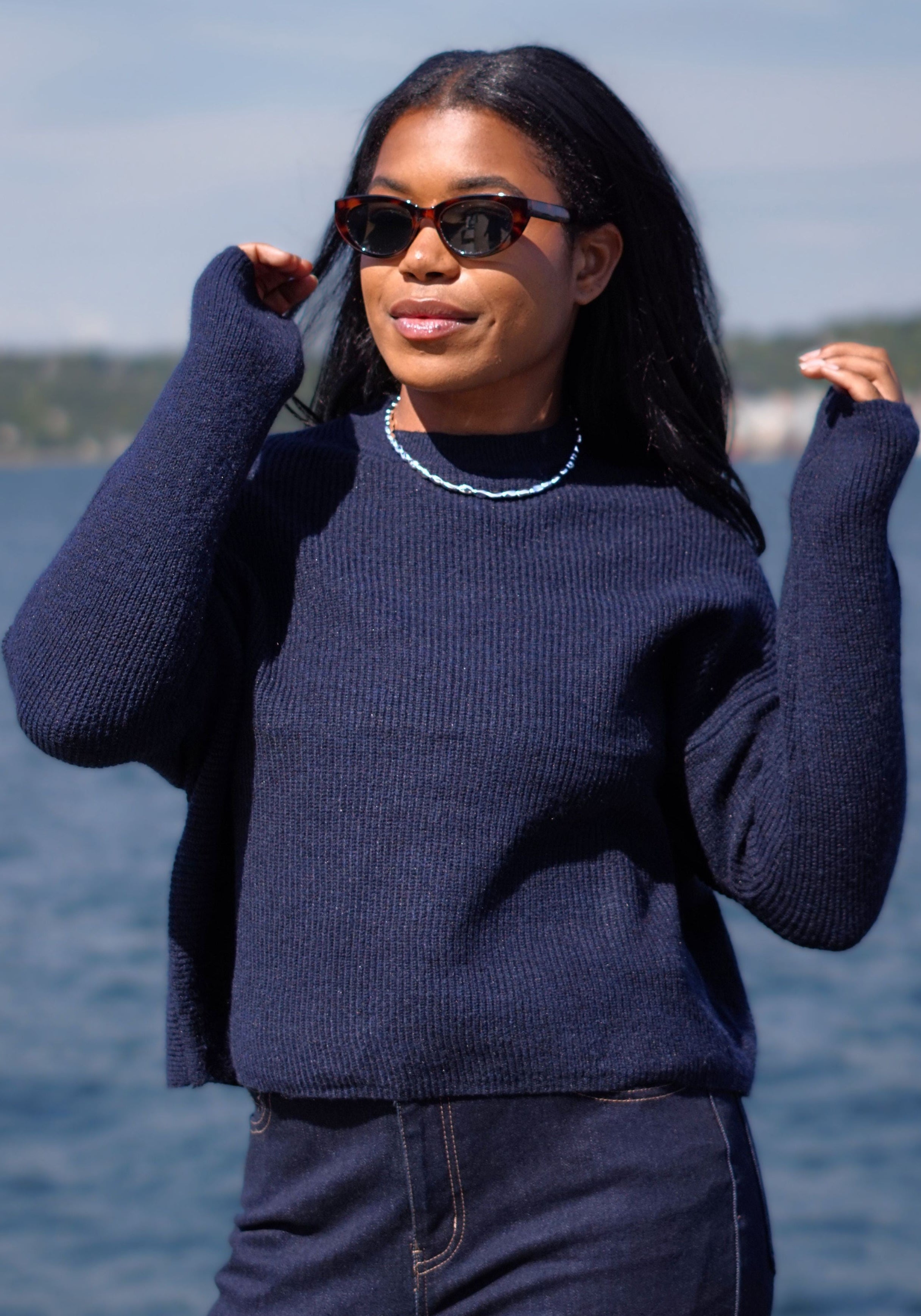 A woman wearing a dark navy blue ribbed knit sweater and matching dark jeans stands outdoors by the water, wearing tortoiseshell sunglasses and a beaded necklace.