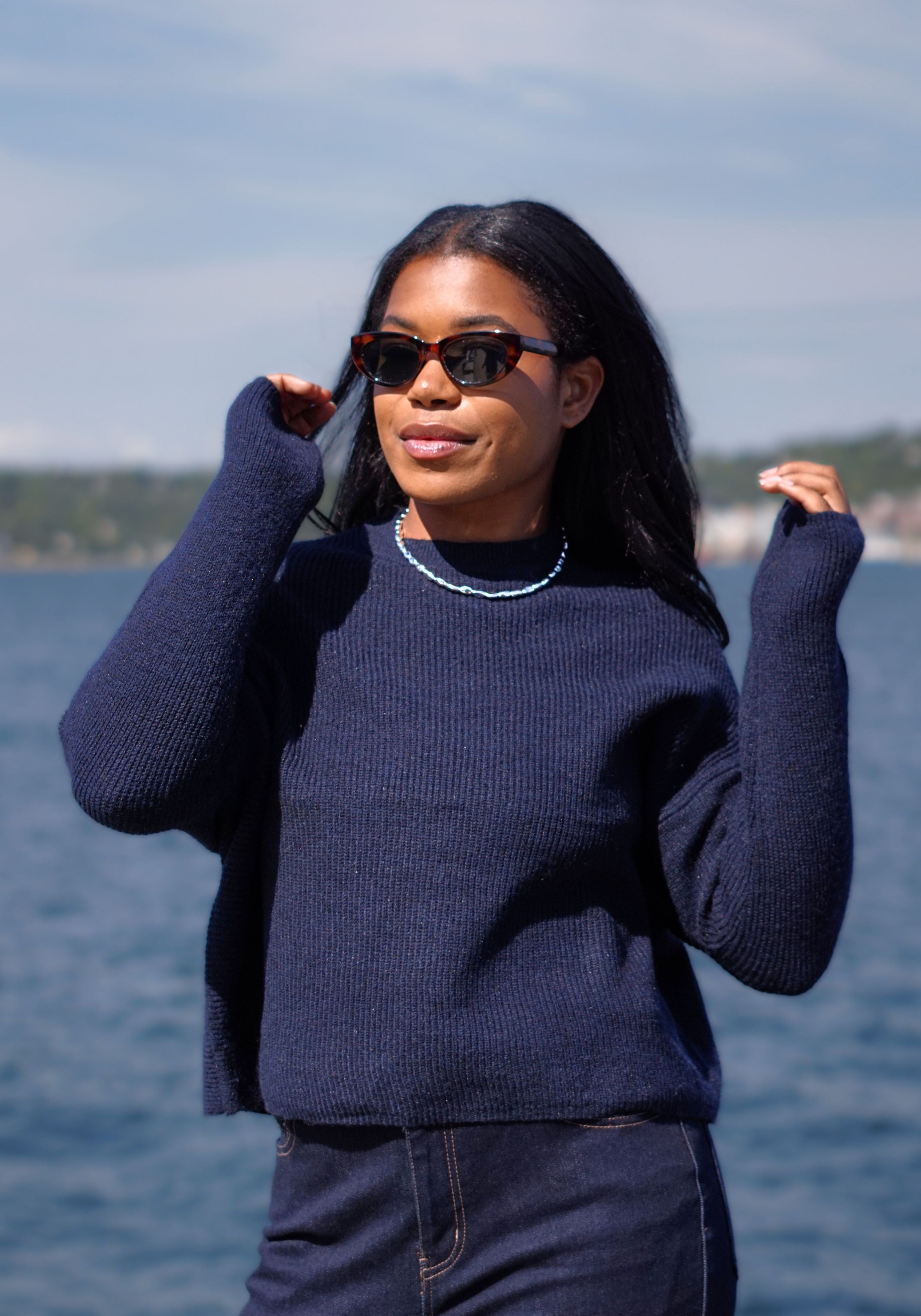 A woman wearing a dark navy blue ribbed knit sweater and matching dark jeans stands outdoors by the water, wearing tortoiseshell sunglasses and a beaded necklace.