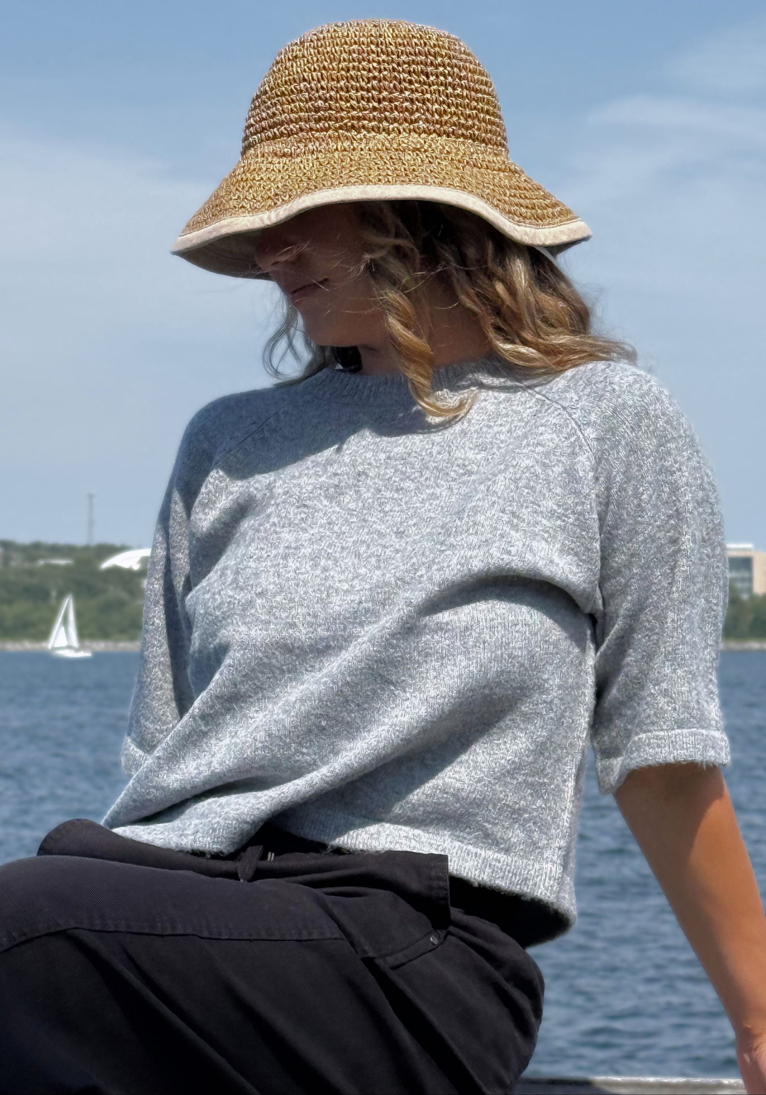 A woman wearing a light gray knit short-sleeve top and a woven straw bucket hat, sitting outdoors.