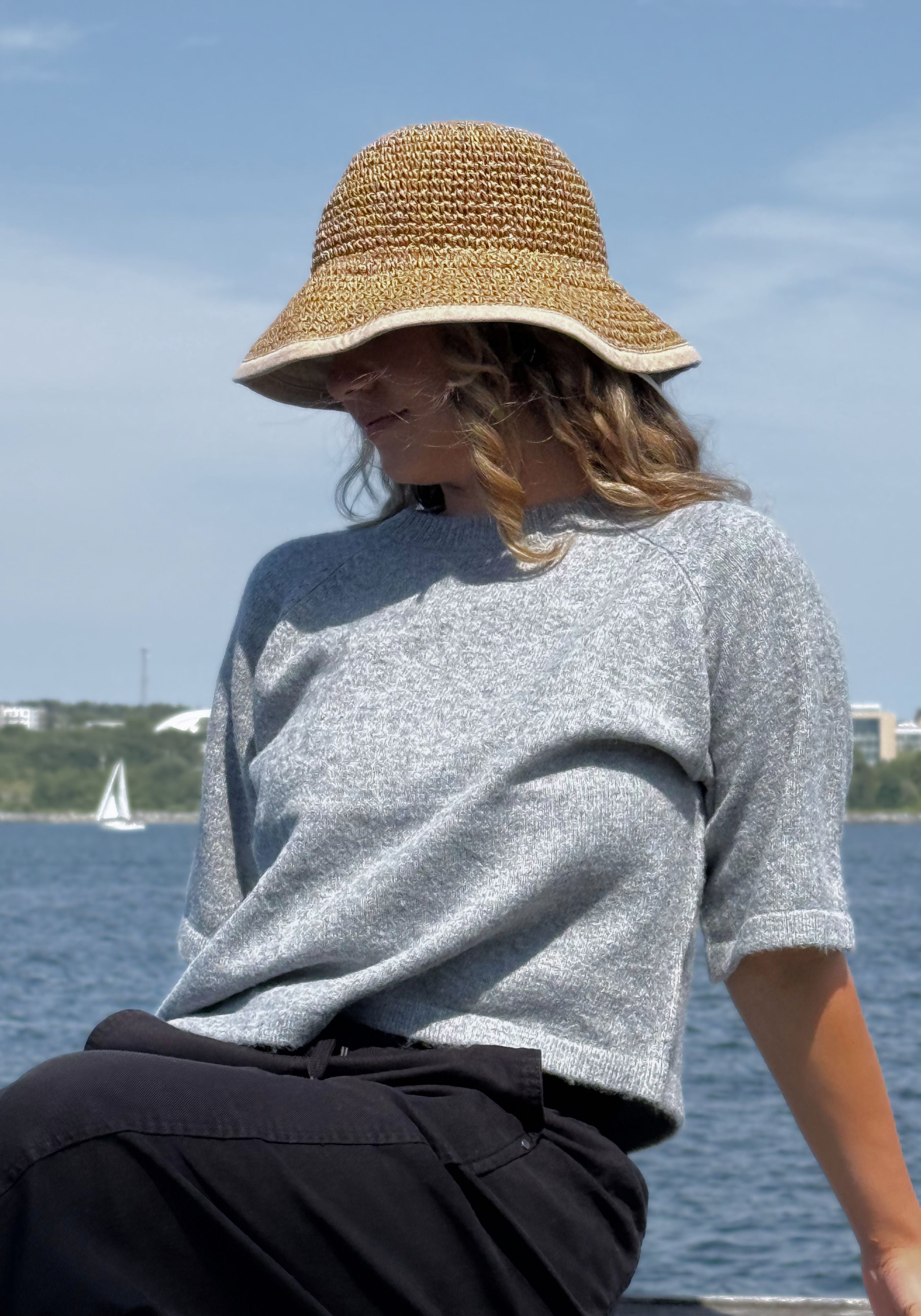 A woman wearing a light gray knit short-sleeve top and a woven straw bucket hat, sitting outdoors.