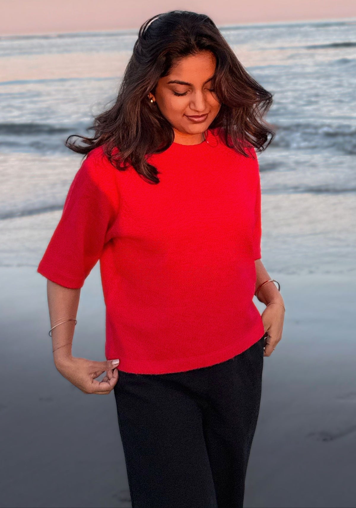 A woman standing on a beach at sunset wearing a bright red short-sleeve top and black pants.