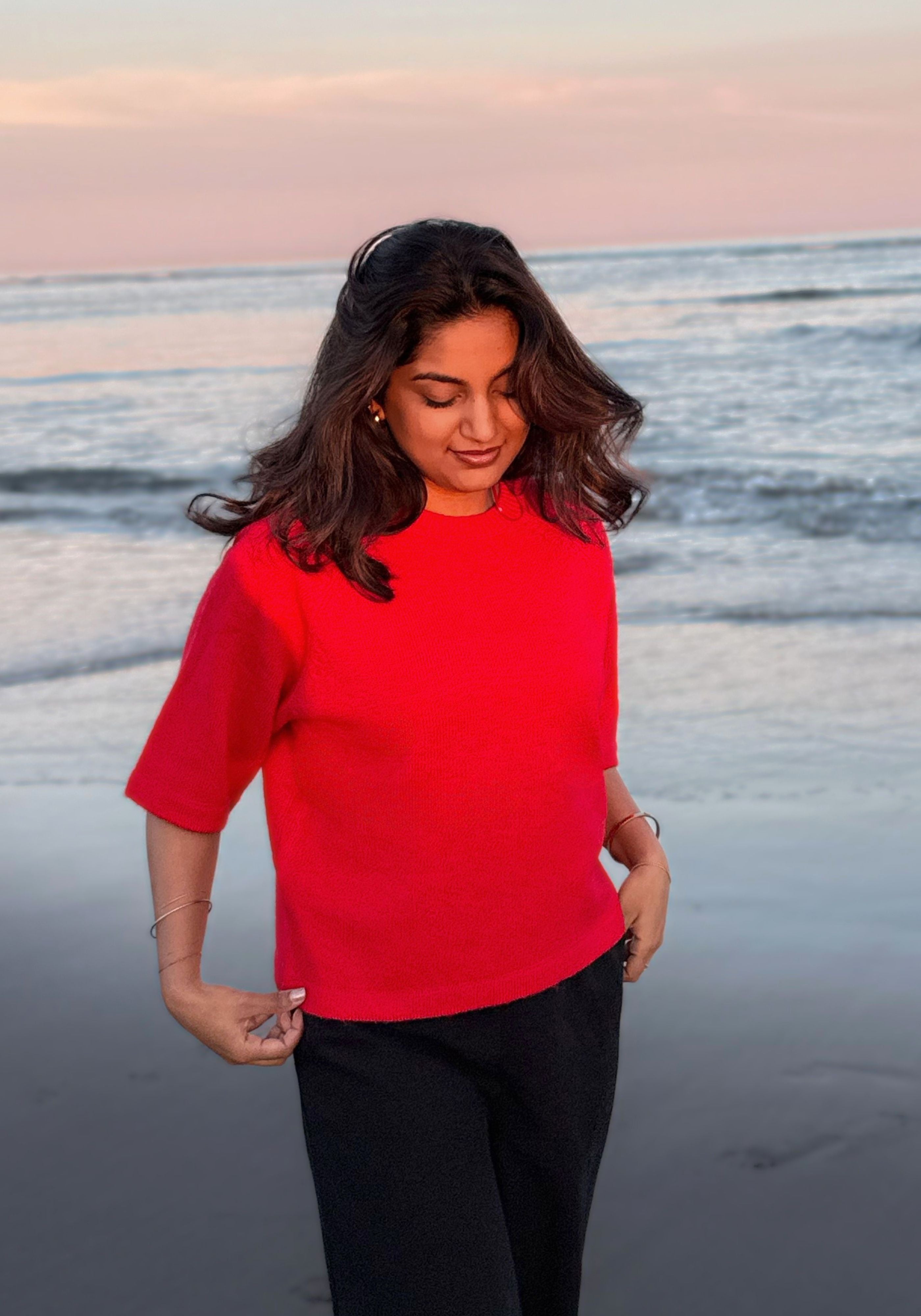 A woman standing on a beach at sunset wearing a bright red short-sleeve top and black pants.
