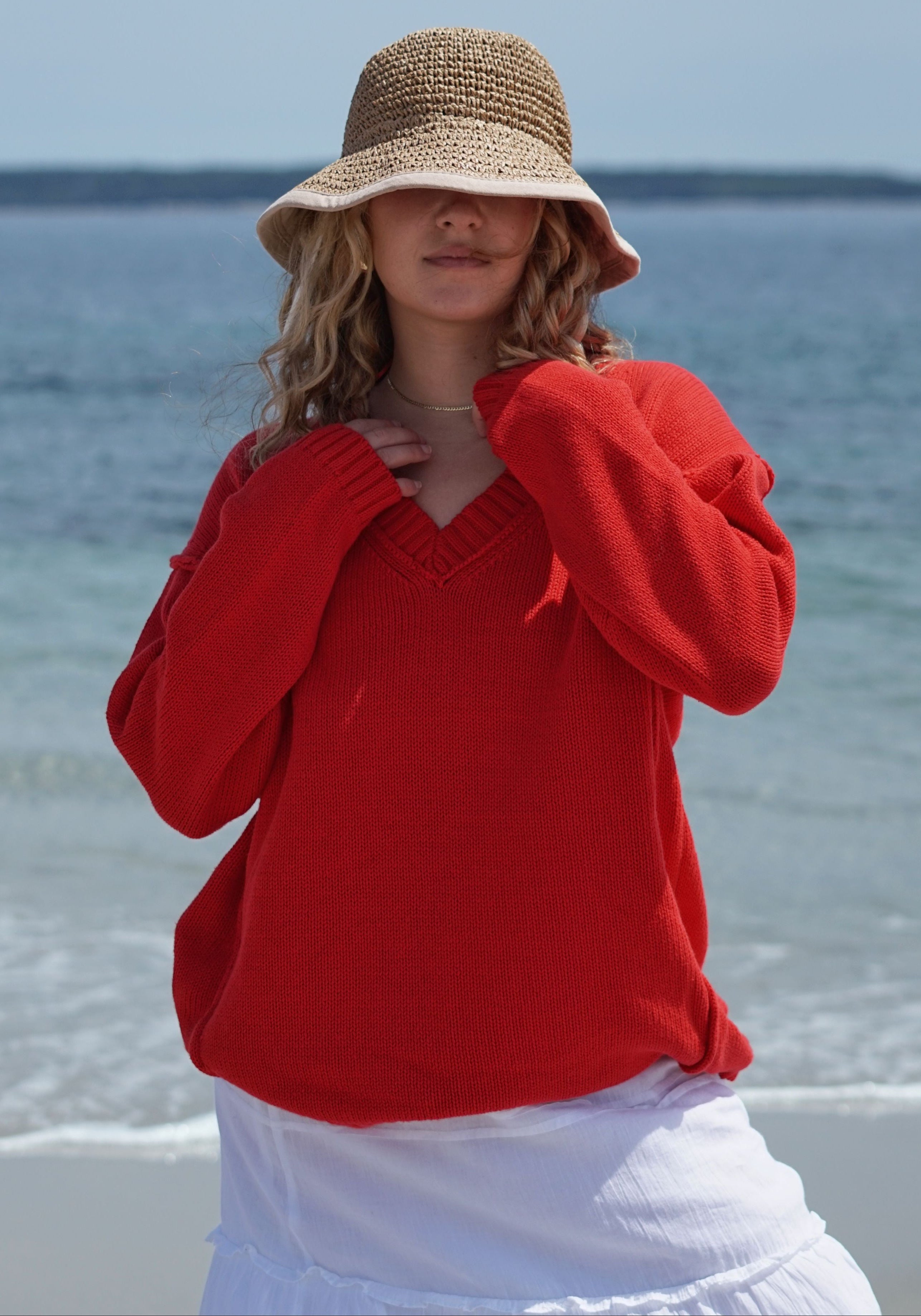 A woman standing on a beach wearing a bright red knit sweater, a straw sun hat, and a white skirt.