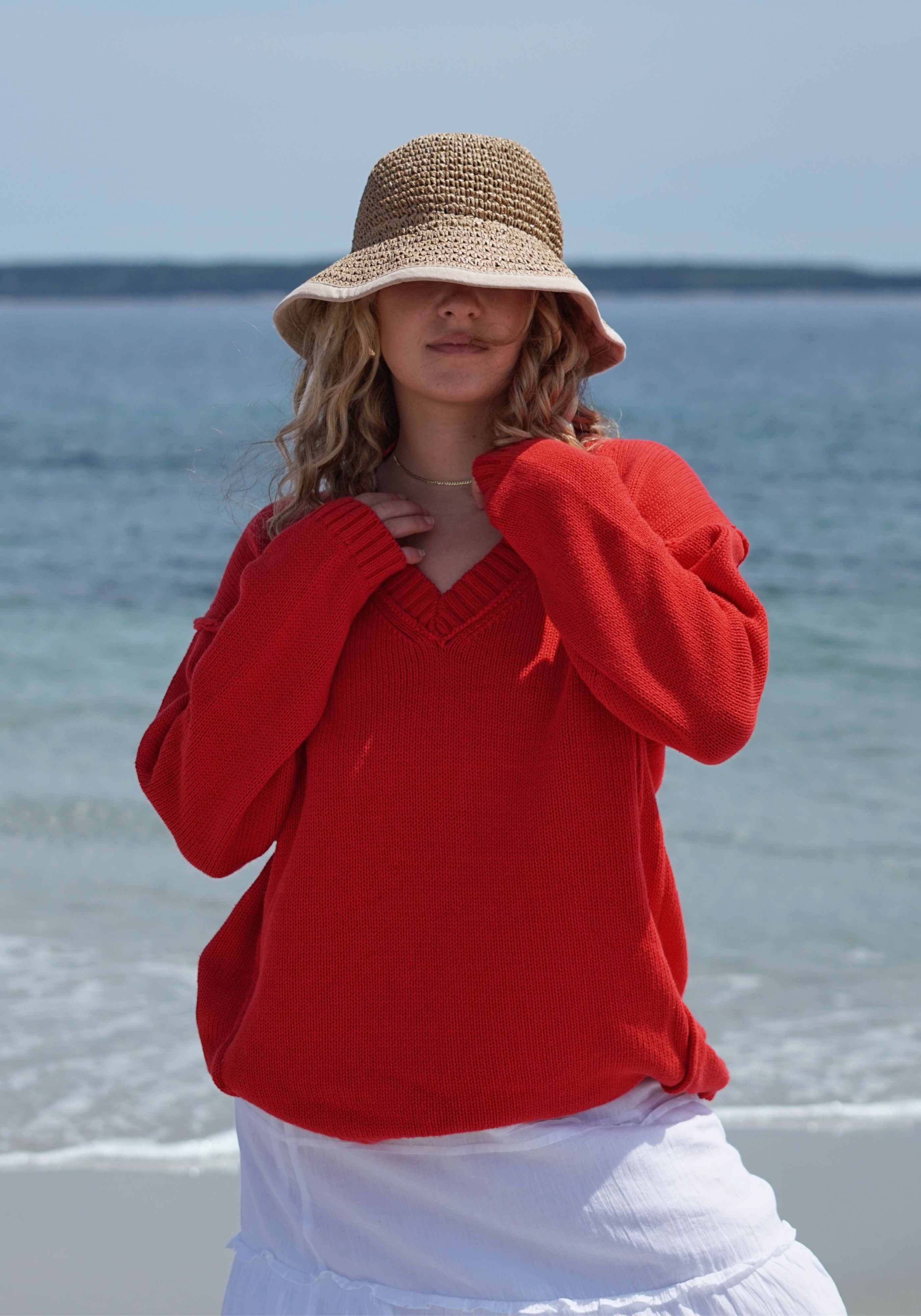 A woman standing on a beach wearing a bright red knit sweater, a straw sun hat, and a white skirt.