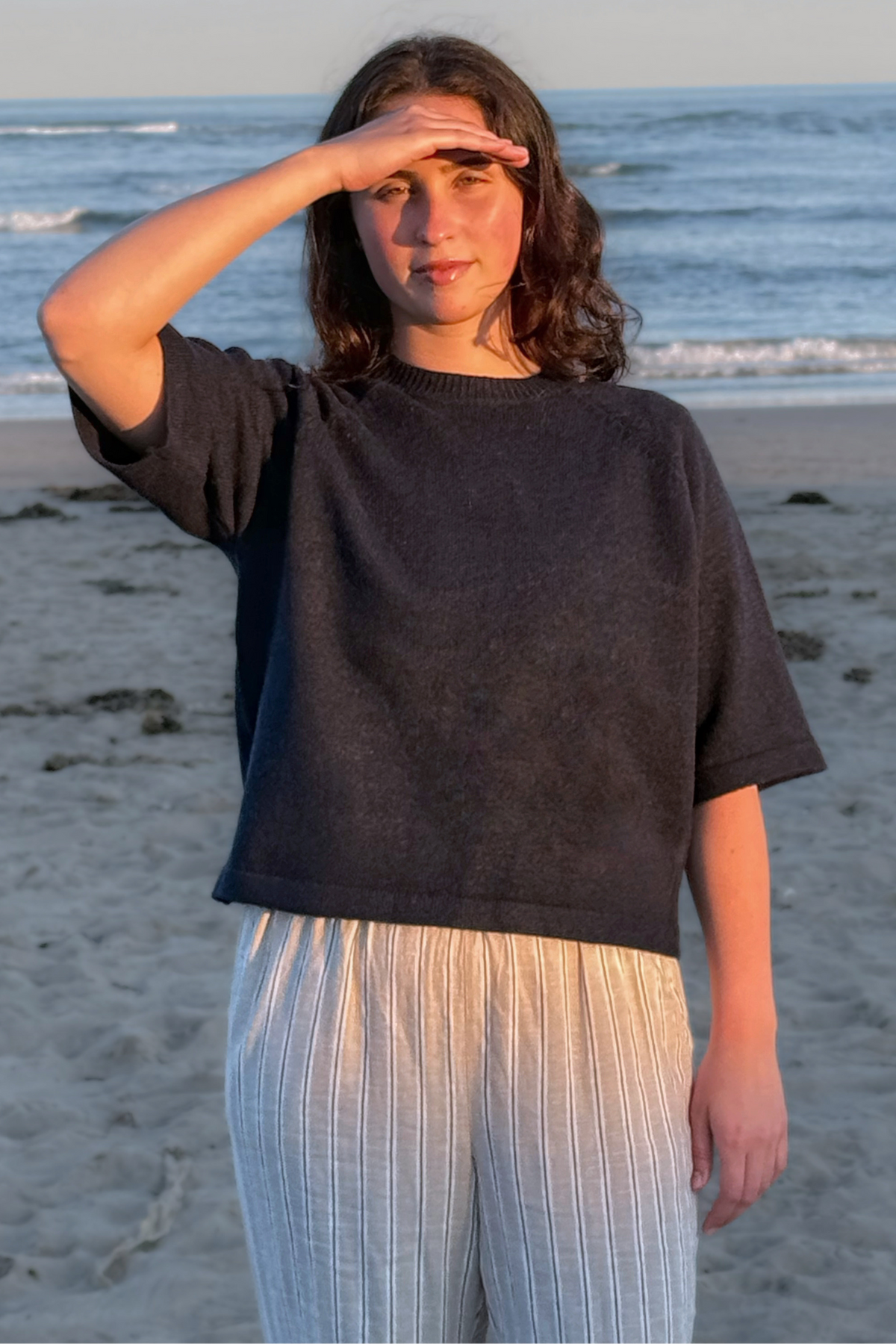 A woman standing on a sandy beach with the ocean in the background, wearing a dark charcoal gray oversized short-sleeve knit top and light beige vertically striped pants.