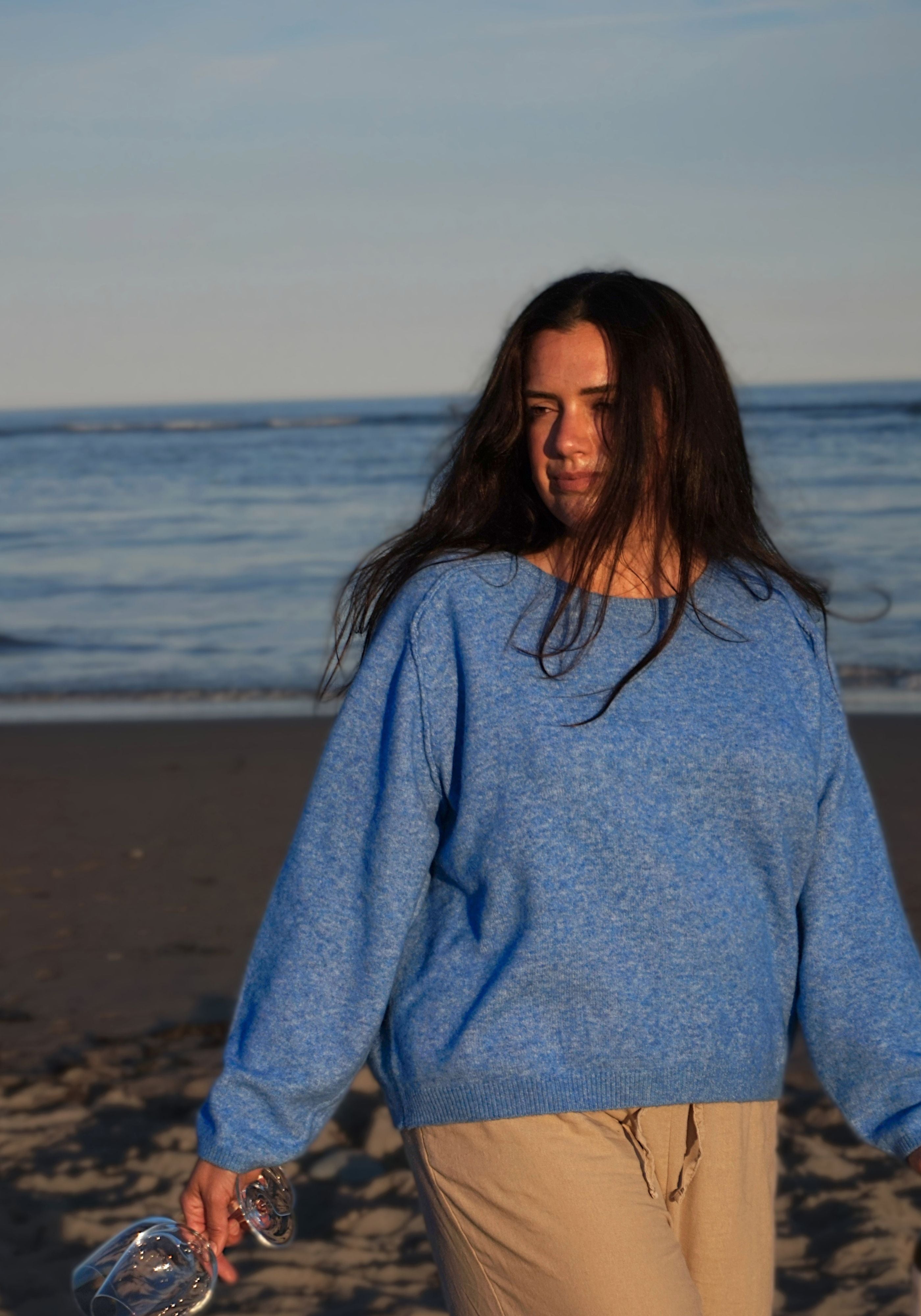 A woman with long dark hair stands on a beach wearing a heathered light blue knit sweater and beige pants, holding wine glasses.