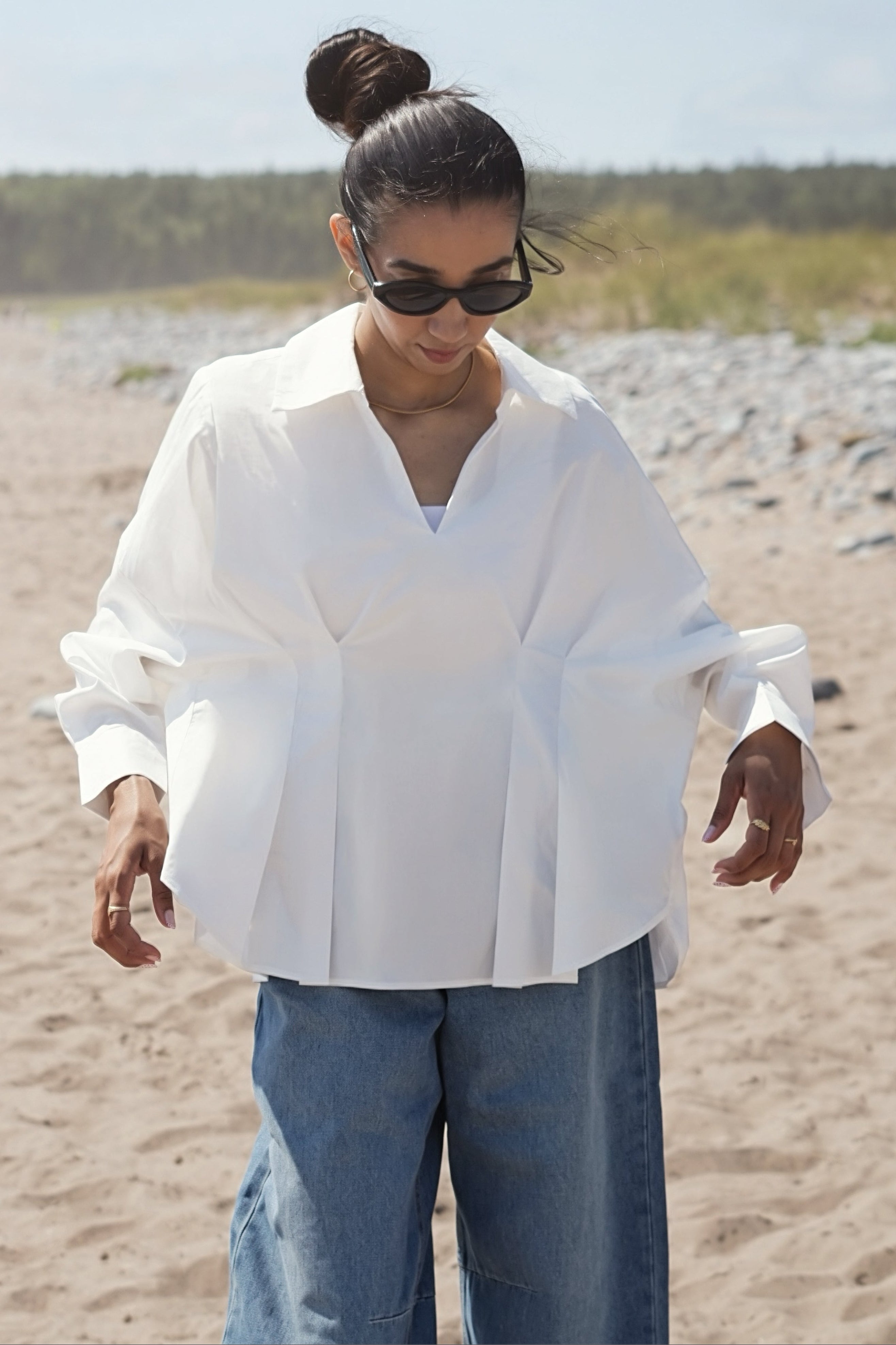 A woman wearing an oversized white long-sleeve blouse and blue jeans walking on a sandy beach.