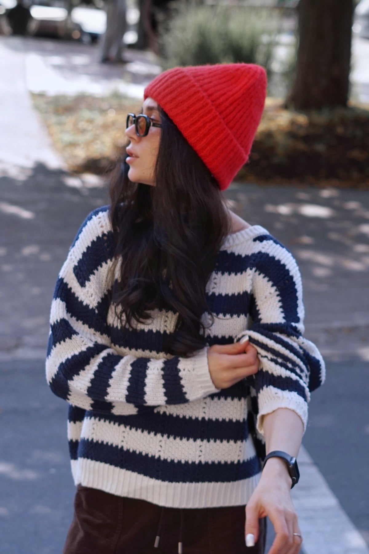 A woman wearing a red knit beanie, a navy and cream striped knit sweater, and brown pants, standing outdoors on a paved path.