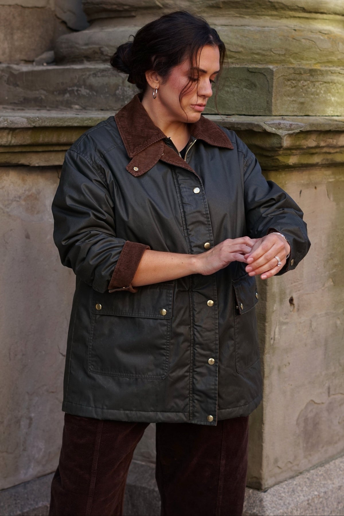 A woman wearing a dark olive green waxed cotton jacket with a brown corduroy collar and cuffs, standing outdoors against a stone wall.