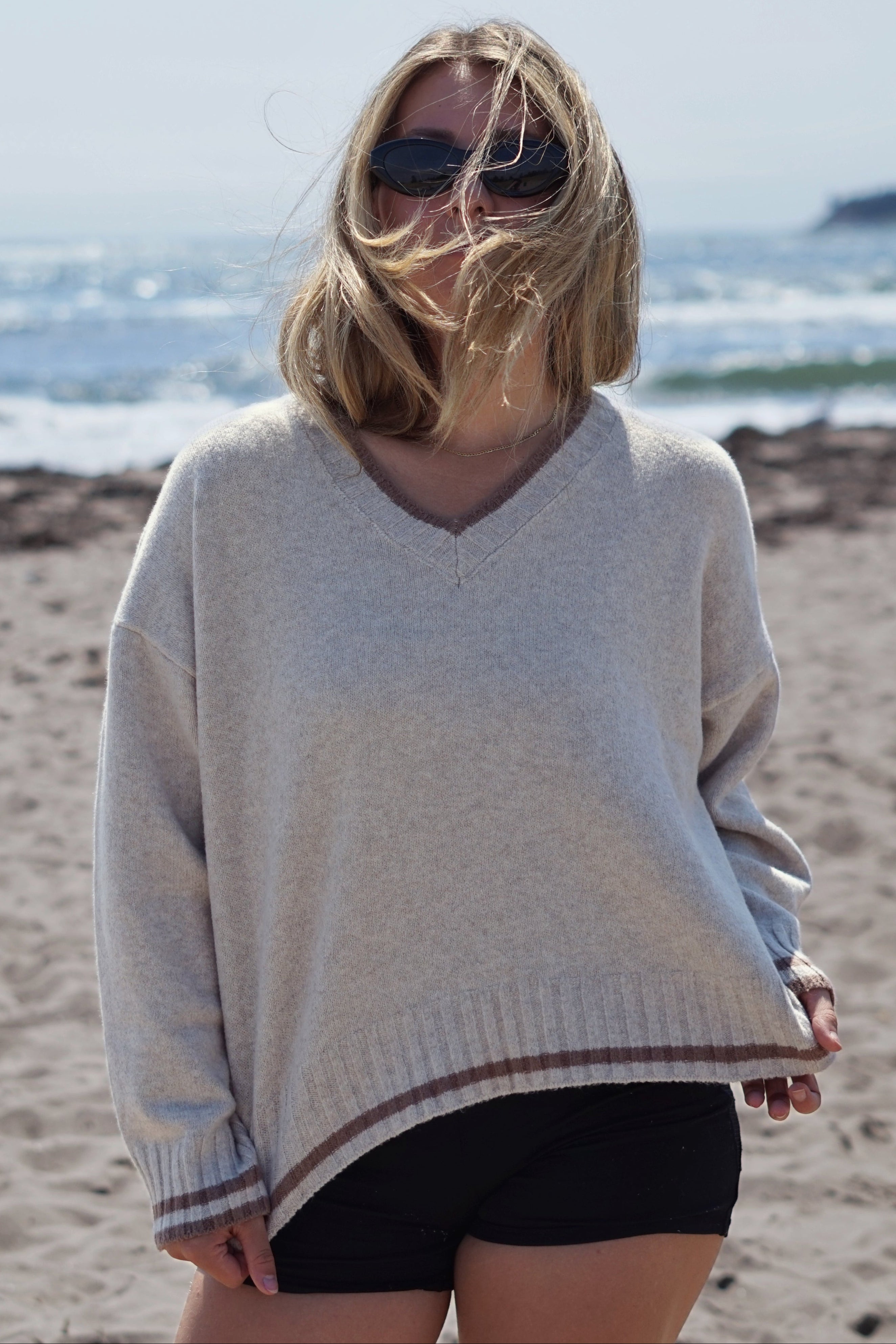 A woman standing on a sandy beach wearing an oversized light beige knit sweater with brown and white striped ribbed trim, black shorts, and dark sunglasses, with the ocean in the background.