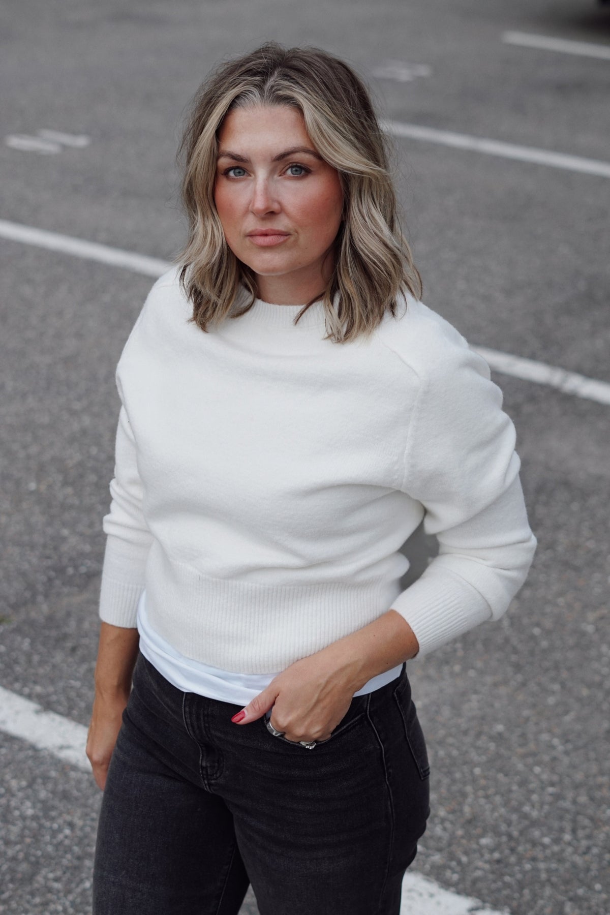 A woman with blonde-highlighted shoulder-length hair wearing a white ribbed mock-neck sweater and black jeans, standing in an outdoor paved parking lot.