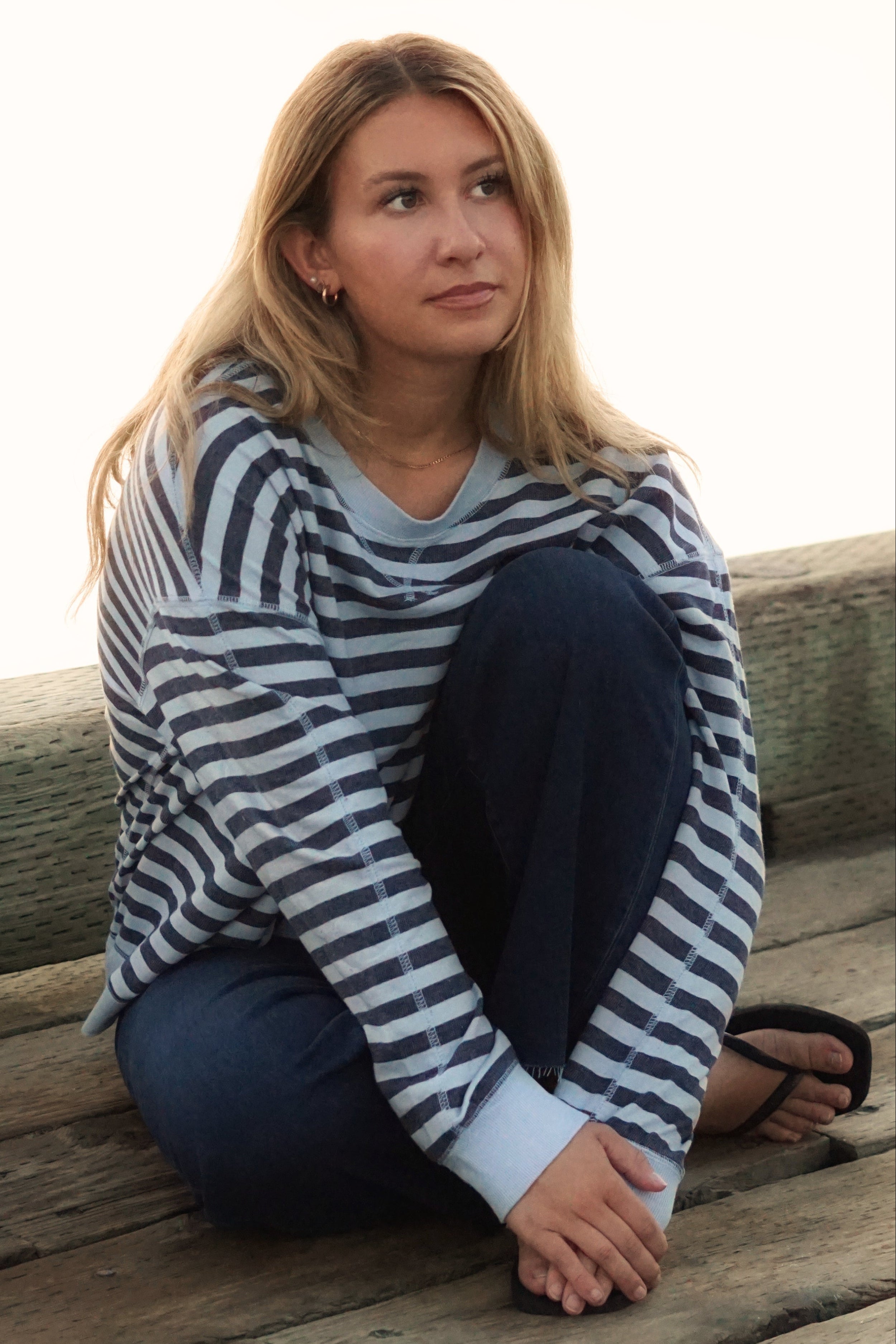 A woman sits on a weathered wooden dock wearing a navy and white horizontally striped long-sleeve shirt, dark blue jeans, and black sandals, with a bright, overexposed outdoor background.