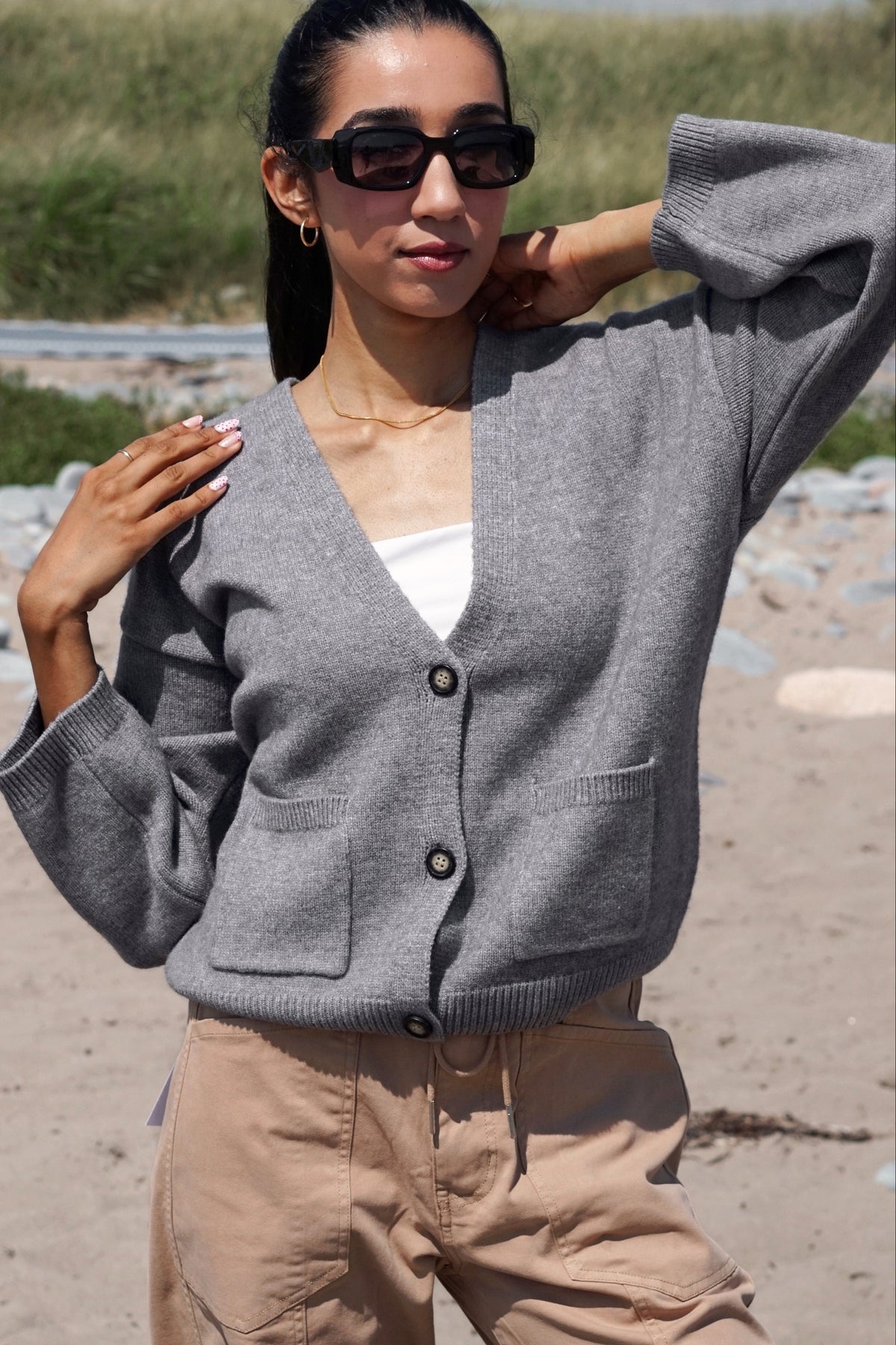 A woman wearing a gray knit cardigan with front buttons, beige pants, and black sunglasses, standing on a sandy beach with grassy dunes in the background.