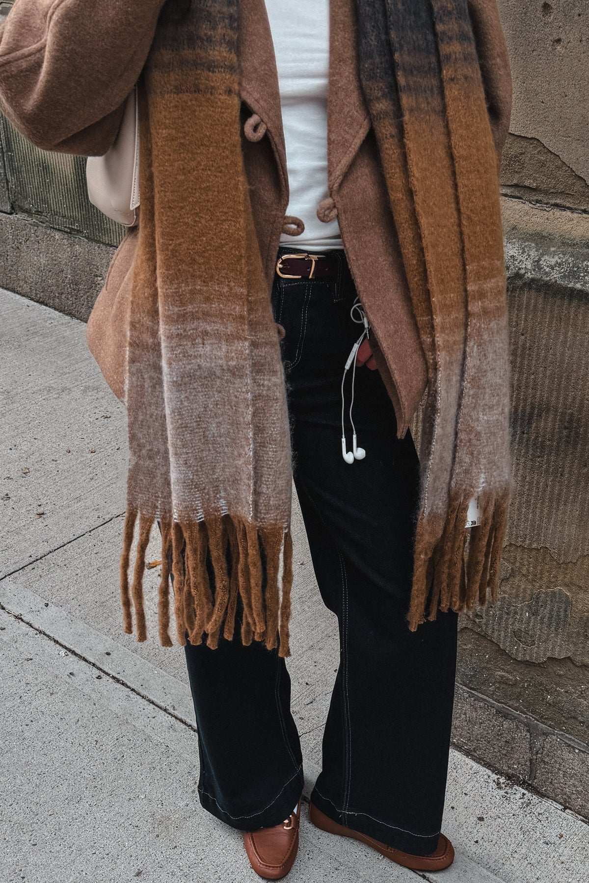 A person standing outdoors on a sidewalk wearing a brown and beige fringed scarf, a tan cardigan, a white top, black wide-leg pants, and brown shoes.