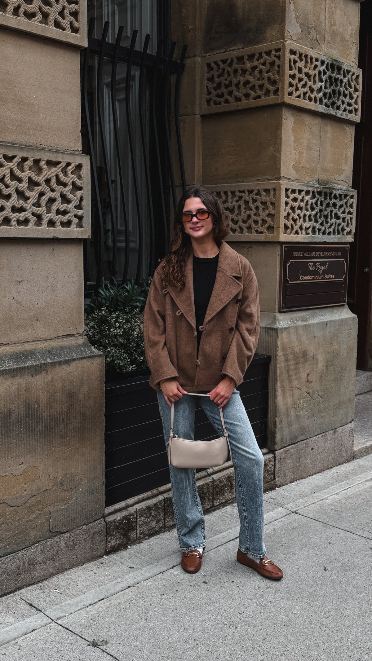 A woman stands on a city sidewalk wearing a brown suede jacket, light blue jeans, brown leather loafers, and holding a small beige handbag. She is posed in front of a stone building facade with decorative stone blocks and a black metal window.