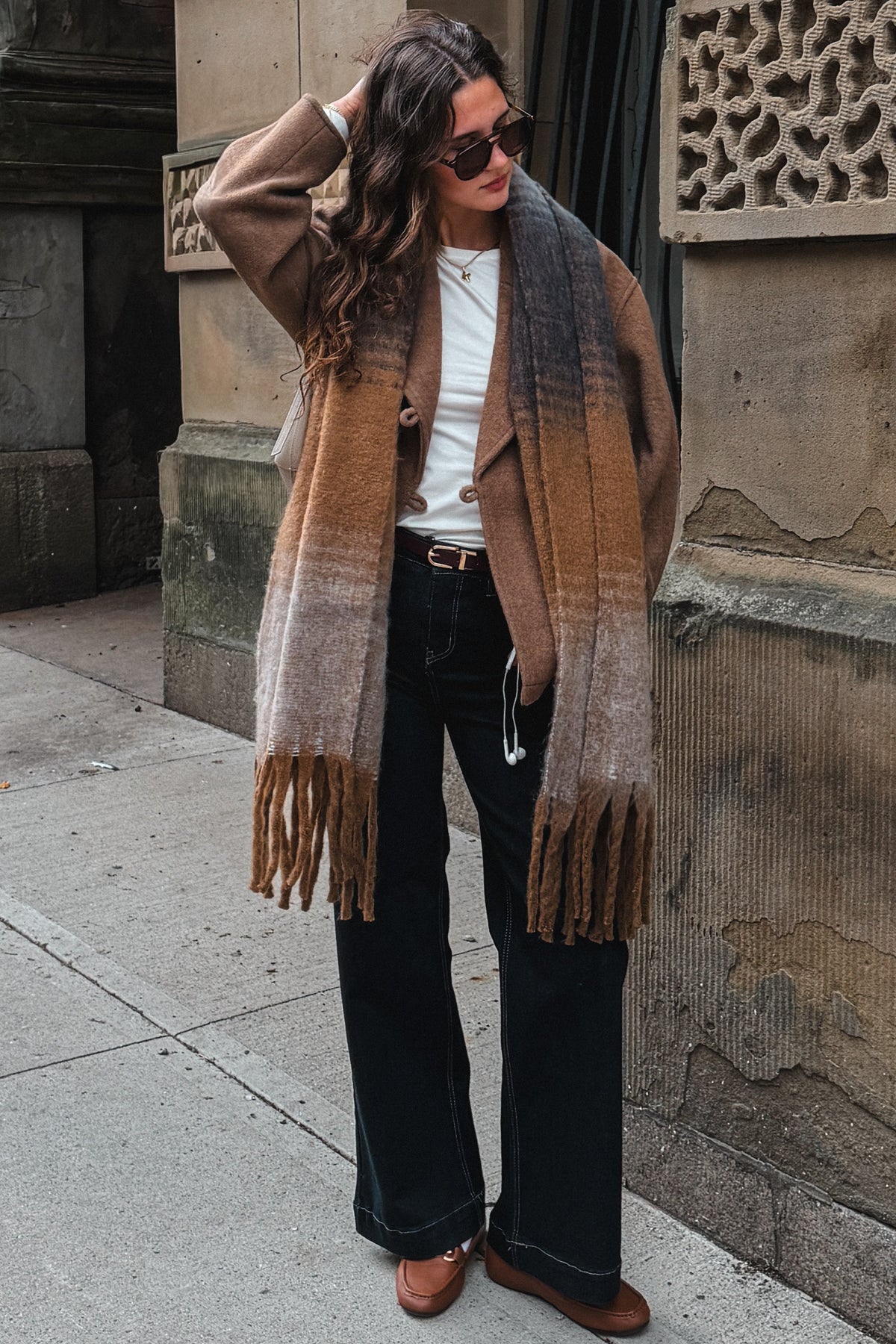 A woman stands on a city sidewalk wearing a long brown and beige ombre fringed scarf draped over her shoulders, a white top, black wide-leg pants, and brown loafers, posing against a stone building facade.