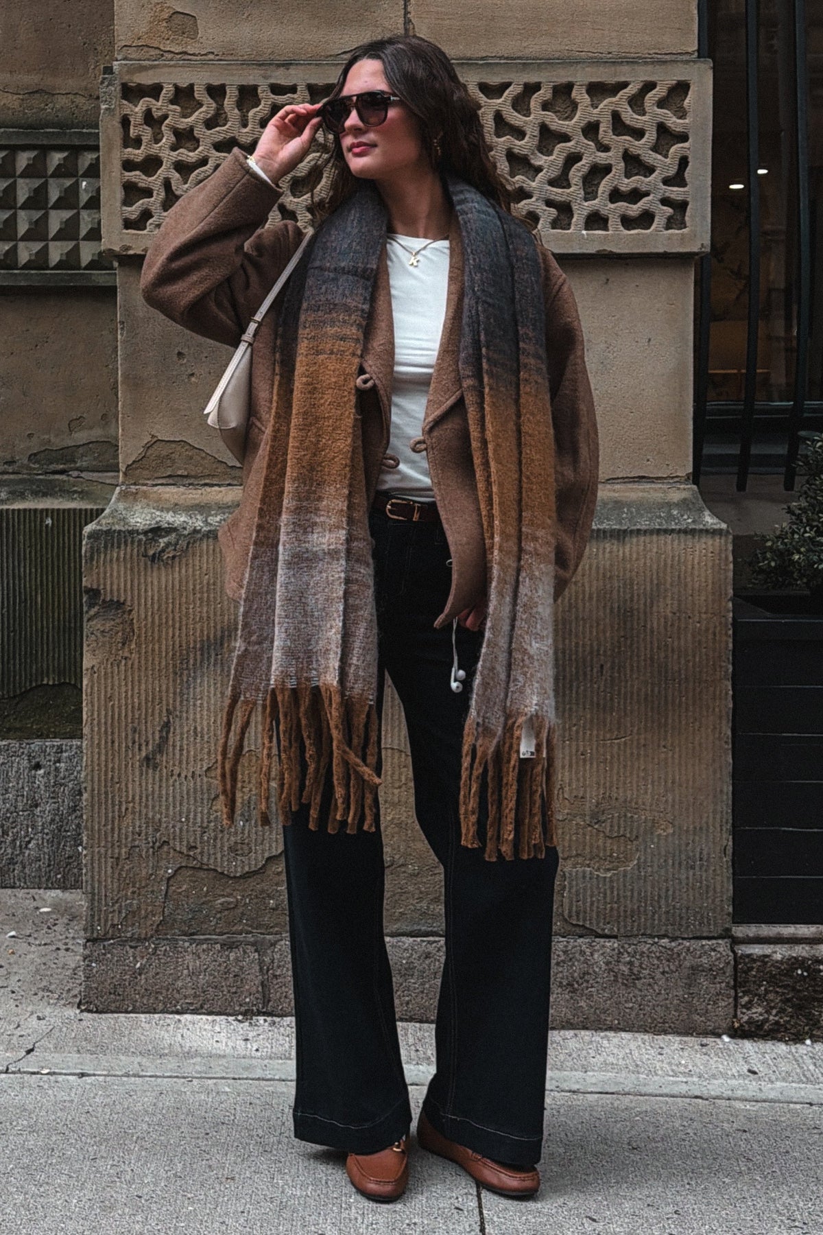 A woman stands on a city sidewalk in front of a stone building, wearing a brown and beige gradient fringed scarf, a brown jacket, a white top, black wide-leg pants, and brown shoes.