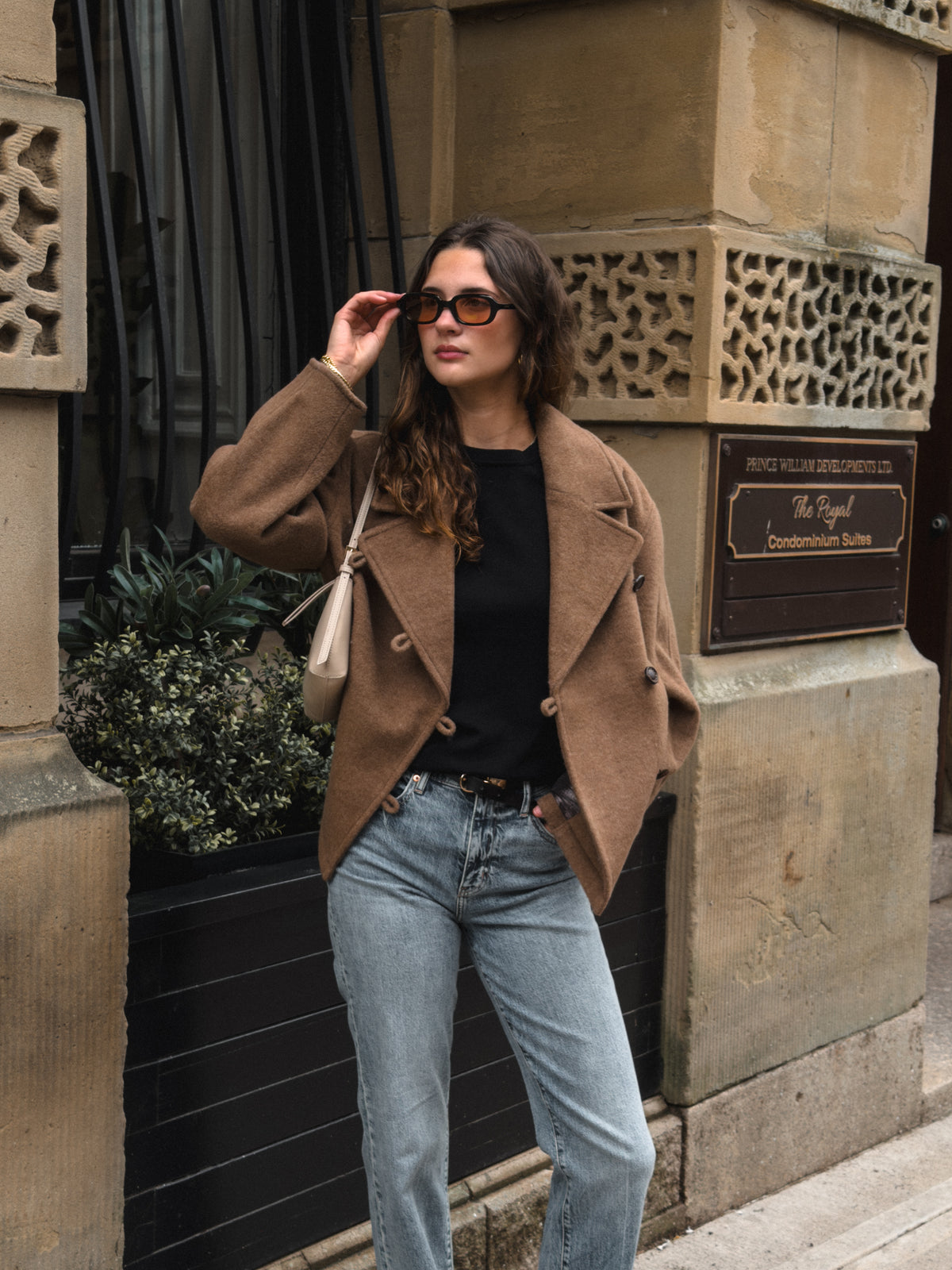 A woman posing outdoors in a brown double-breasted blazer, black top, light-wash blue jeans, and sunglasses, carrying a beige shoulder bag.