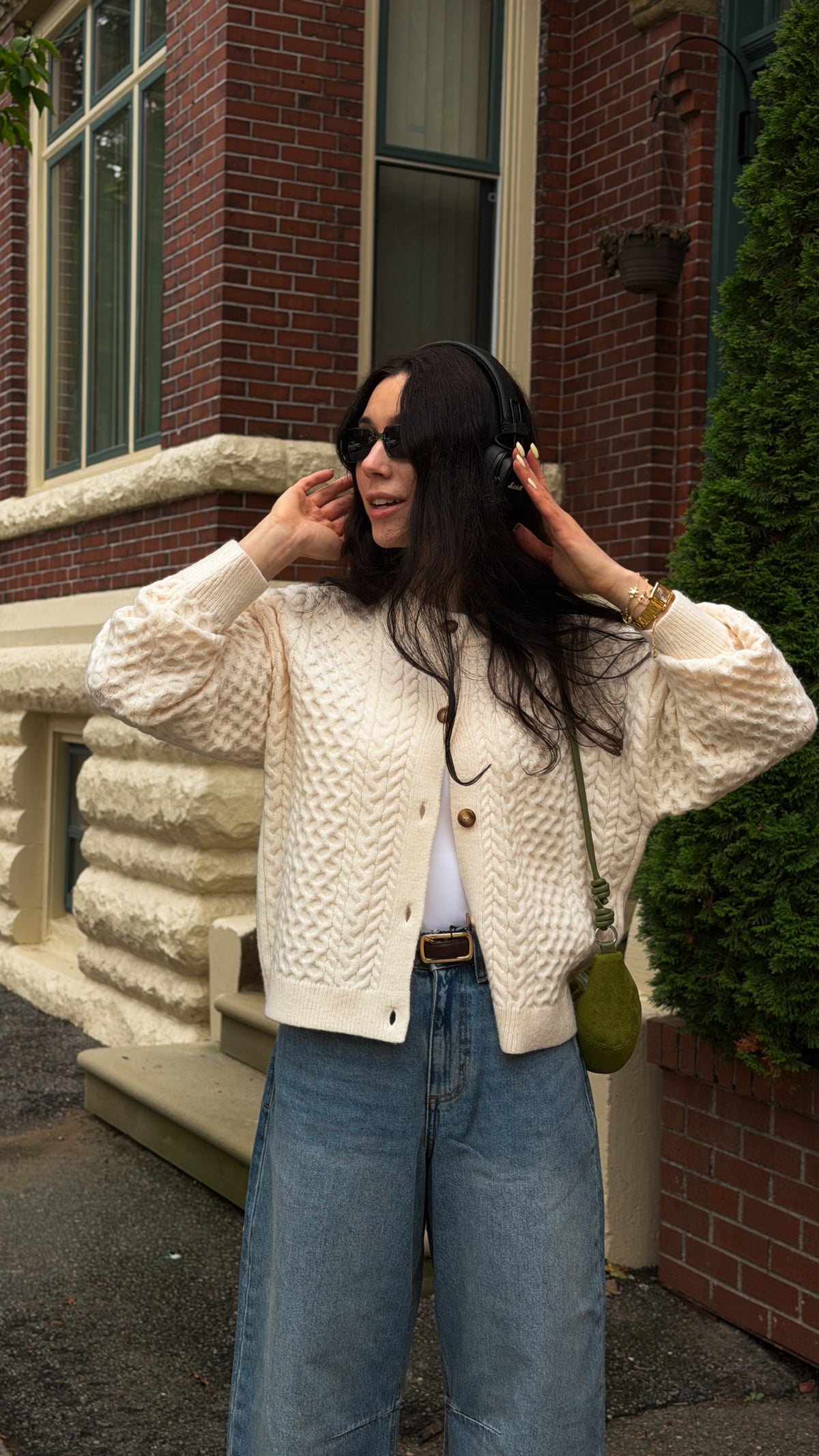 A woman wearing a cream cable-knit cardigan, blue jeans, and black sunglasses stands outdoors in front of a brick building, holding over-ear headphones to her head.