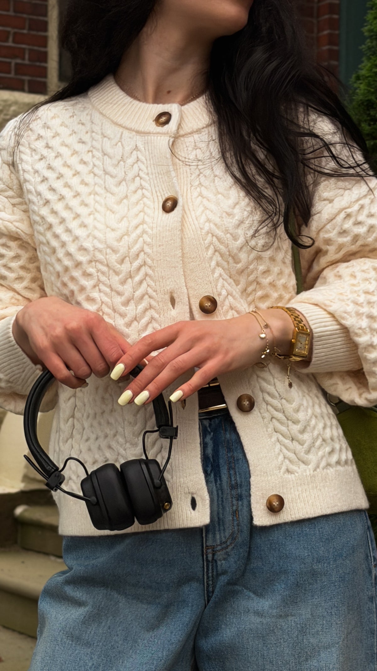 A woman wearing a cream cable-knit cardigan with brown buttons, paired with blue jeans and holding black over-ear headphones.