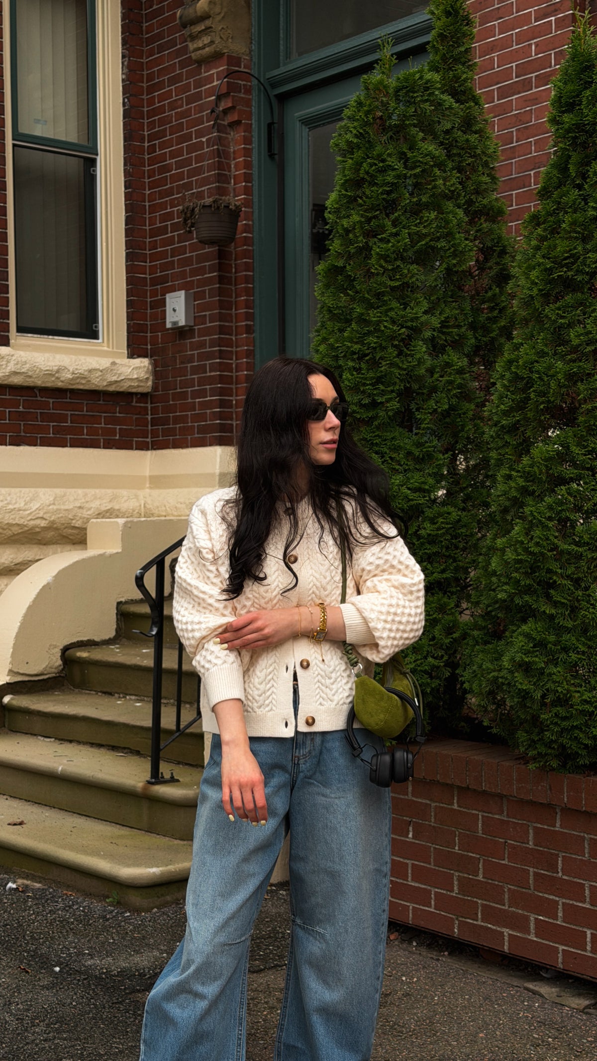 A woman stands outdoors in front of a brick building wearing a cream textured knit cardigan with gold buttons, blue wide-leg jeans, and a small olive green shoulder bag.