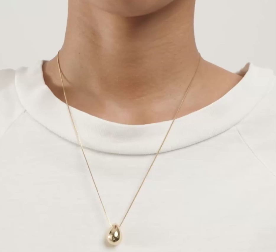 A close-up of a person wearing a delicate gold chain necklace with a small teardrop-shaped gold pendant, styled with a white crew-neck t-shirt.