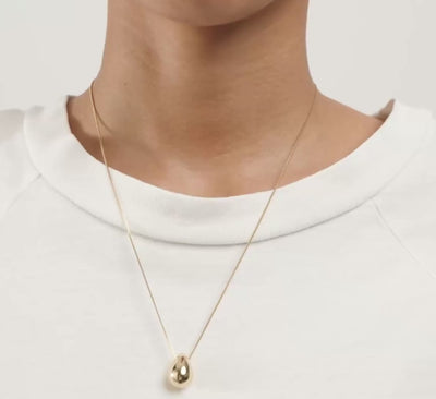A close-up of a person wearing a delicate gold chain necklace with a small teardrop-shaped gold pendant, styled with a white crew-neck t-shirt.