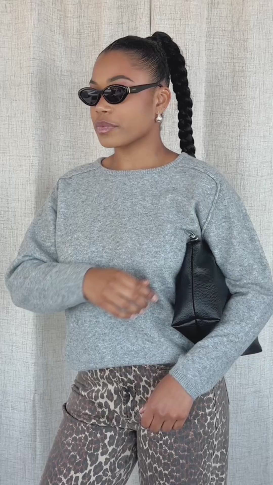A woman modeling a light grey heathered knit sweater and brown leopard-print pants with black sunglasses, standing against a neutral beige fabric backdrop.