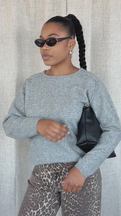 A woman modeling a light grey heathered knit sweater and brown leopard-print pants with black sunglasses, standing against a neutral beige fabric backdrop.