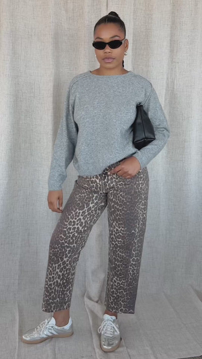 A woman modeling a light gret heathered knit sweater and leopard-print pants against a neutral beige fabric backdrop.