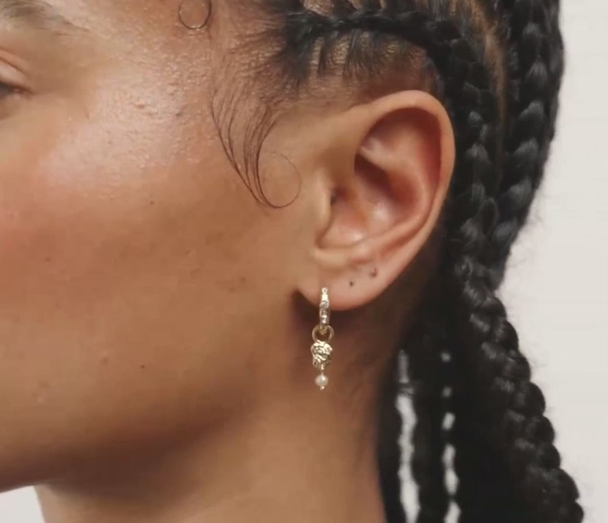 A close-up of a person's ear wearing a small gold huggie hoop earring adorned with clear stones and a dangling pearl.