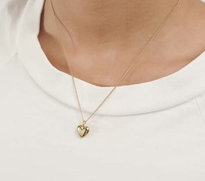 A close-up portrait of a woman with dark hair wearing a delicate gold heart pendant necklace on a dark navy top, set against a dark background.