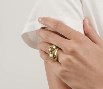 A close-up lifestyle shot of a model wearing a chunky gold ring with two stacked, rounded gold discs on her finger, set against a white tee and a light background.