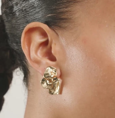 A close-up of a model's ear wearing a gold-toned, organic-shaped stud earring with a small clear stone accent, set against a plain light gray background.